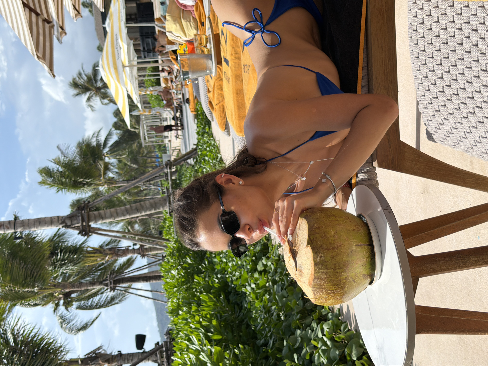 A woman in a blue bikini is drinking from a coconut with a straw while lying on a lounge chair in Choeng Thale, Thailand. She wears sunglasses and has jewelry on her wrist and neck. The coconut sits on a white table next to her lounge chair. Palm trees and other greenery are visible in the background, along with other beachgoers and umbrellas.