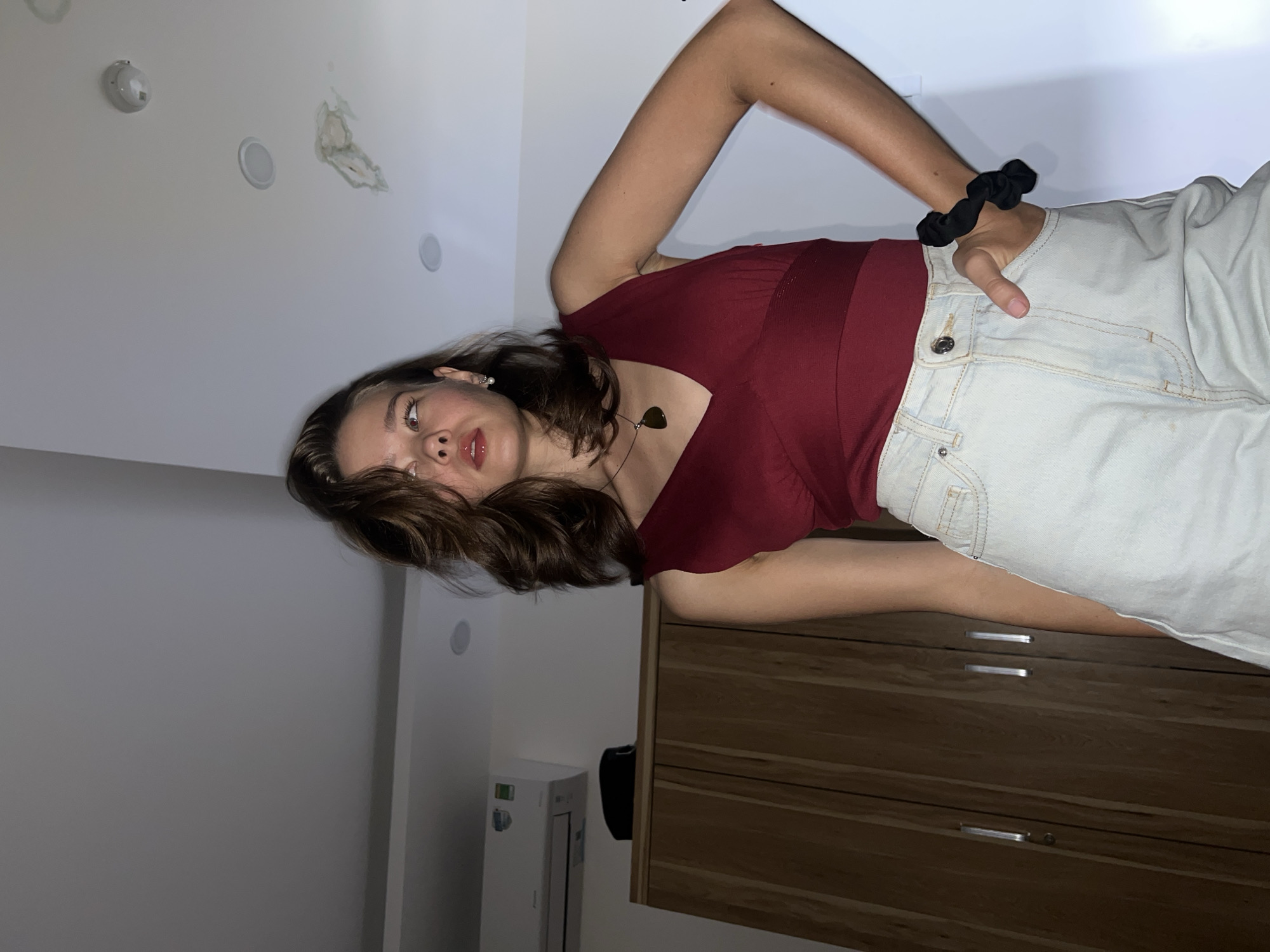 A woman is posing in a room with light grey walls and a white ceiling. The ceiling has a smoke detector, a recessed light, and a patch of peeling paint. She is wearing a dark red sleeveless top, light blue jeans, and a black scrunchie on her wrist. She has brown hair, and is wearing a necklace with a dark stone pendant. She is positioned in front of a wooden dresser with horizontal grain and metal pulls. There is a white air conditioning unit visible in the background. The photo may have been taken indoors at a location in Nam Nha Trang Ward, Vietnam.