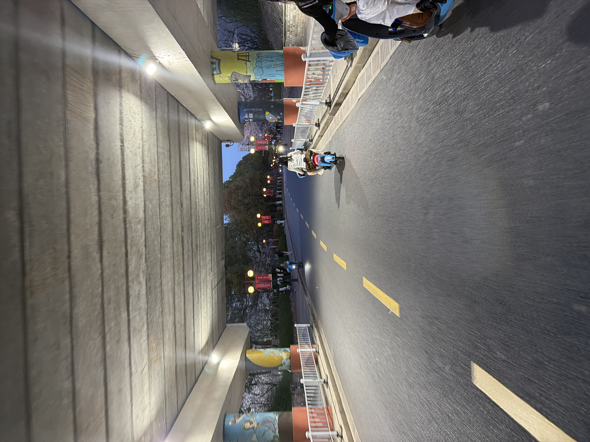 The image depicts a road in Jiangning District, China, likely a bike path, running under a bridge. Yellow lines mark the lane. People are riding what appear to be toy cars or motorized scooters. The bridge supports are decorated with artwork. Lights illuminate the underside of the bridge. The scene seems to be at dusk or early evening. There are trees visible on the side of the road and banners hanging.