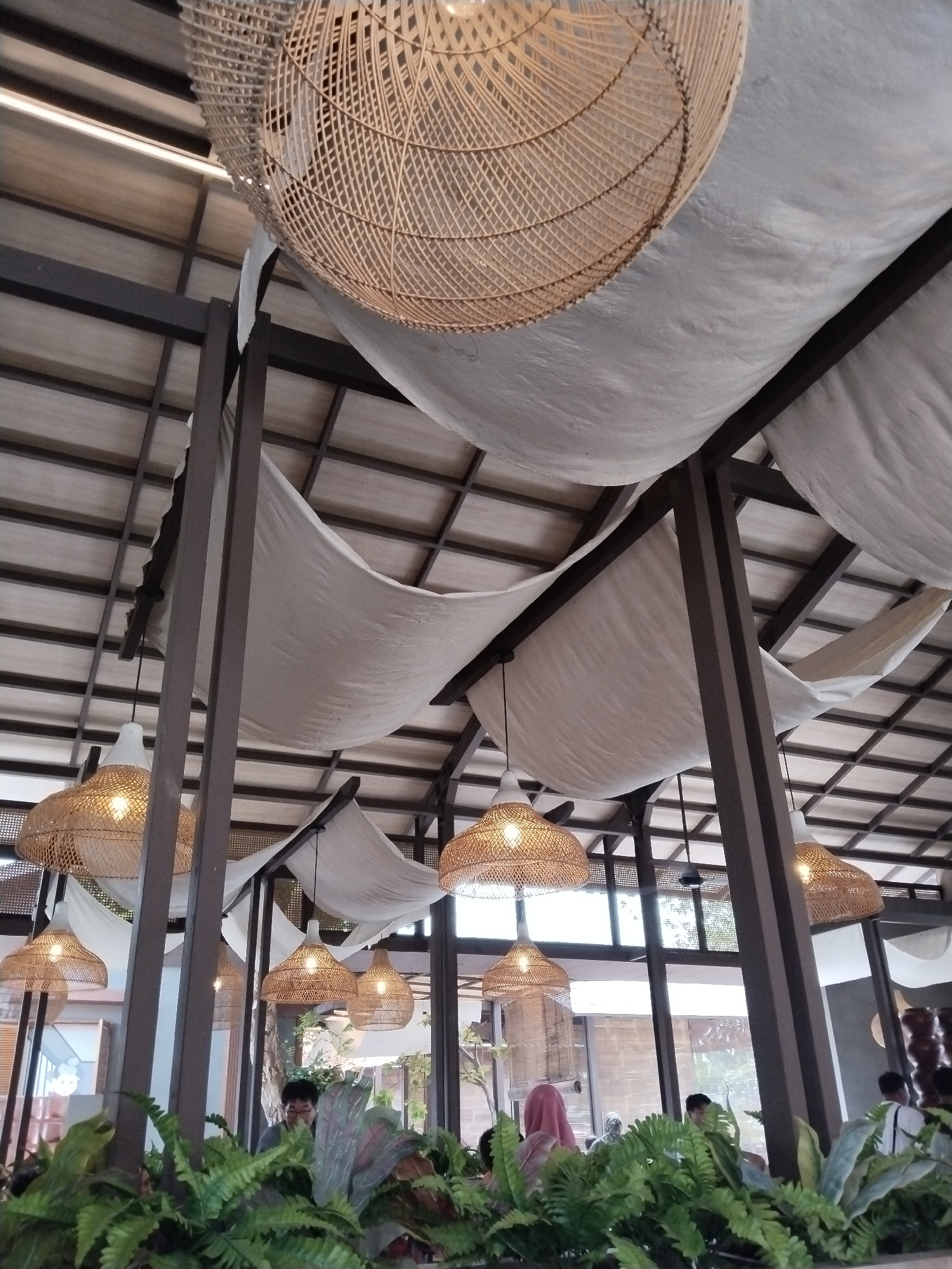 The image depicts the interior of a building, possibly a restaurant or cafe, characterized by a rustic and airy design. The ceiling is adorned with multiple woven rattan pendant lights of various sizes, creating a warm ambiance. Draped white fabric panels are suspended from the ceiling, softening the industrial look of the exposed wooden beams and dark metal supports. In the foreground, lush green potted plants, including ferns, are arranged, suggesting a natural and refreshing atmosphere. Several people are visible in the background, some seated and others standing, engaged in conversations or observing their surroundings. The lighting in the scene is a mix of natural light filtering through windows and the illumination from the pendant lamps. Based on the context provided, this scene is located in Kecamatan Pati, Indonesia.