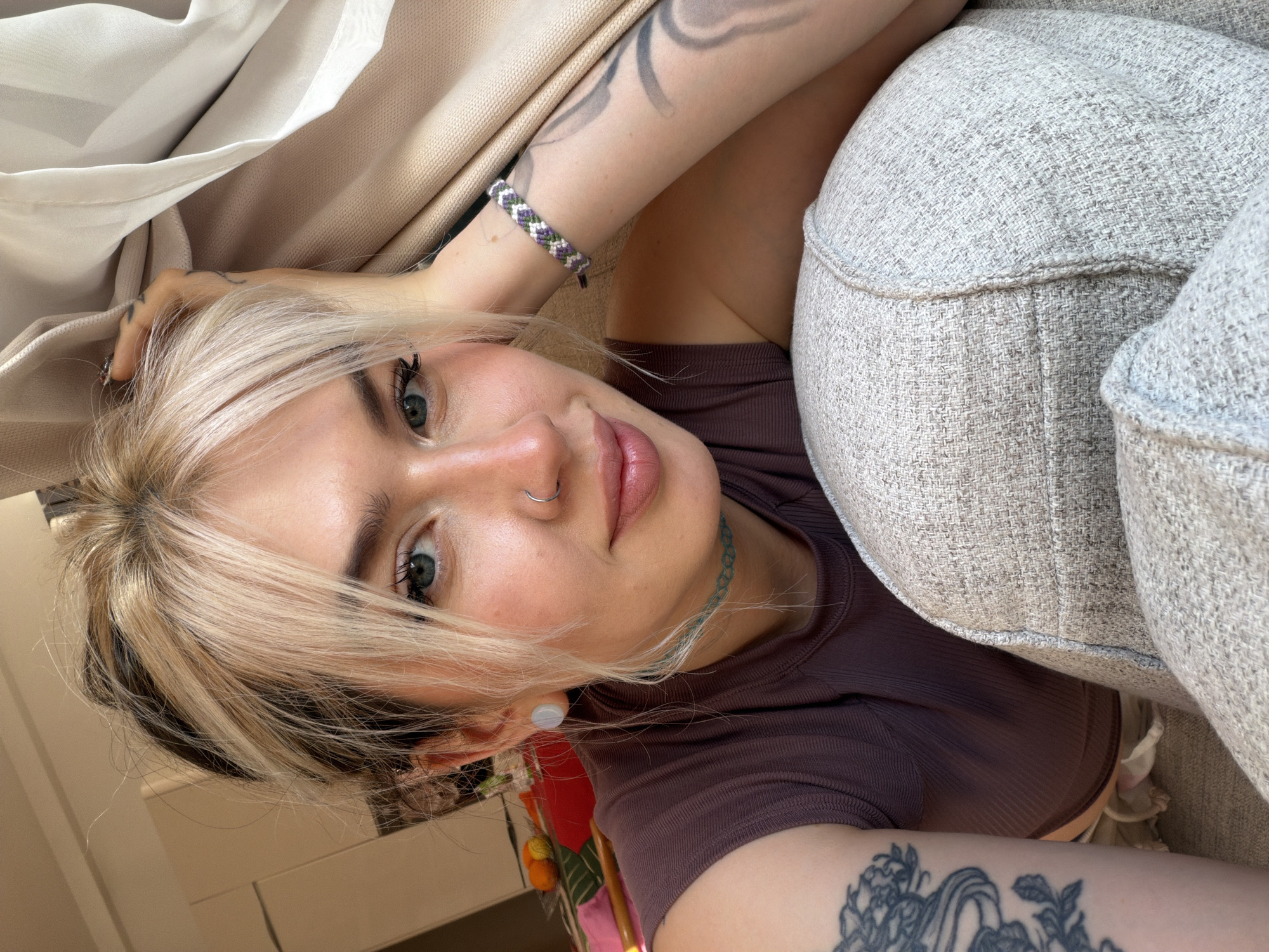 A woman is lying on a light grey upholstered sofa. She is fair-skinned, with blonde hair that has dark roots. She is wearing a brown ribbed shirt, a light blue choker, and a silver nose ring. She has tattoos on both arms and is wearing a bracelet on one wrist. The sofa is light grey with a textured fabric. Behind the sofa is a beige wall or curtain. The location of the image is unknown and not necessarily in Budva, Montenegro. The location of the image can't be derived from the picture alone.