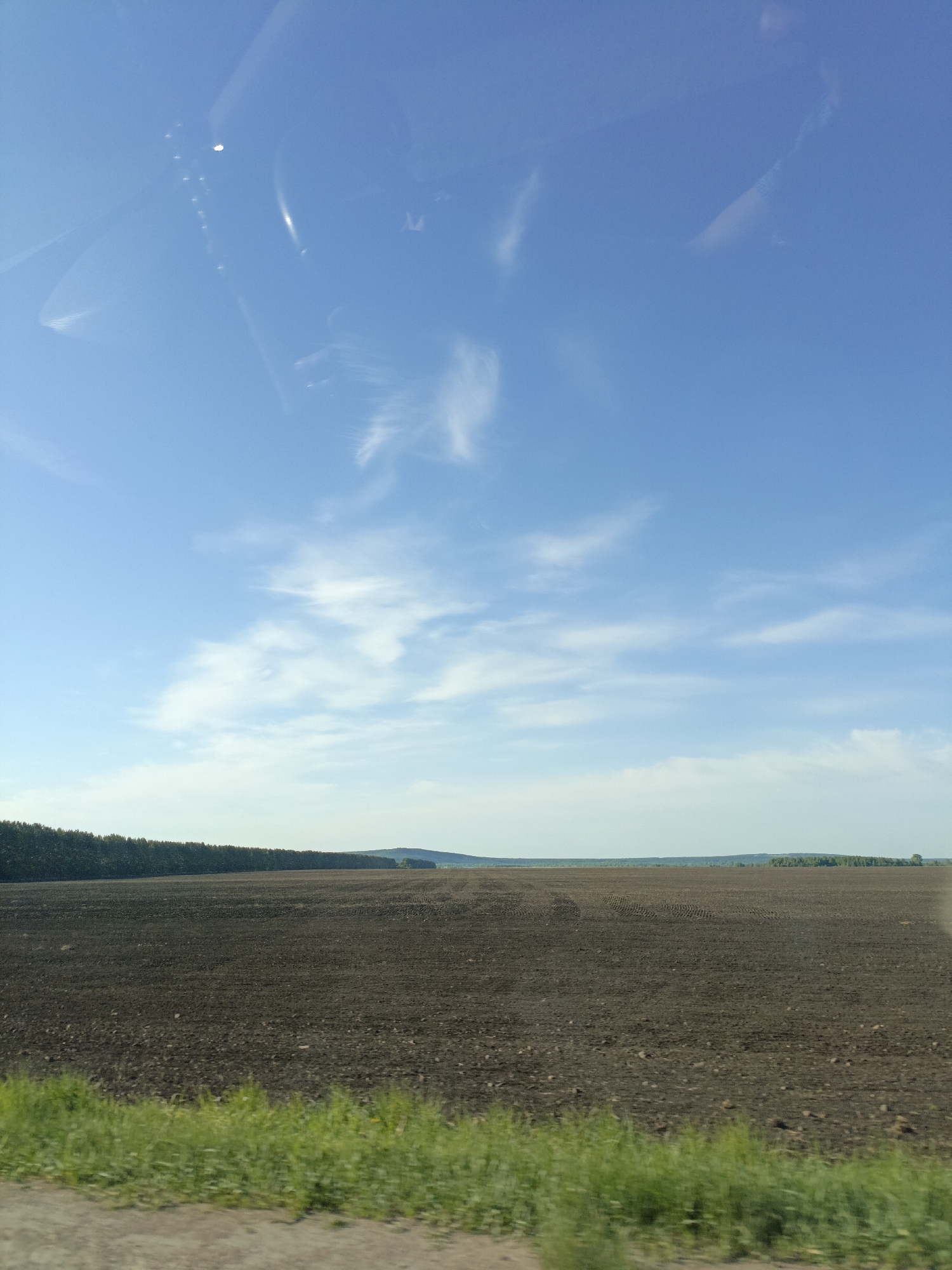 The image depicts a rural landscape in the vicinity of Novoustinovka, Russia. A large, plowed field of dark soil dominates the foreground. In the distance, a line of trees stretches across the horizon, punctuated by a few small hills or rises in the terrain. The sky above is blue with scattered white clouds. The photograph was taken from inside a vehicle, evident from the reflections on the windshield at the top of the frame. A strip of green vegetation is visible along the bottom edge of the image. There are no people present in the visible scene.
