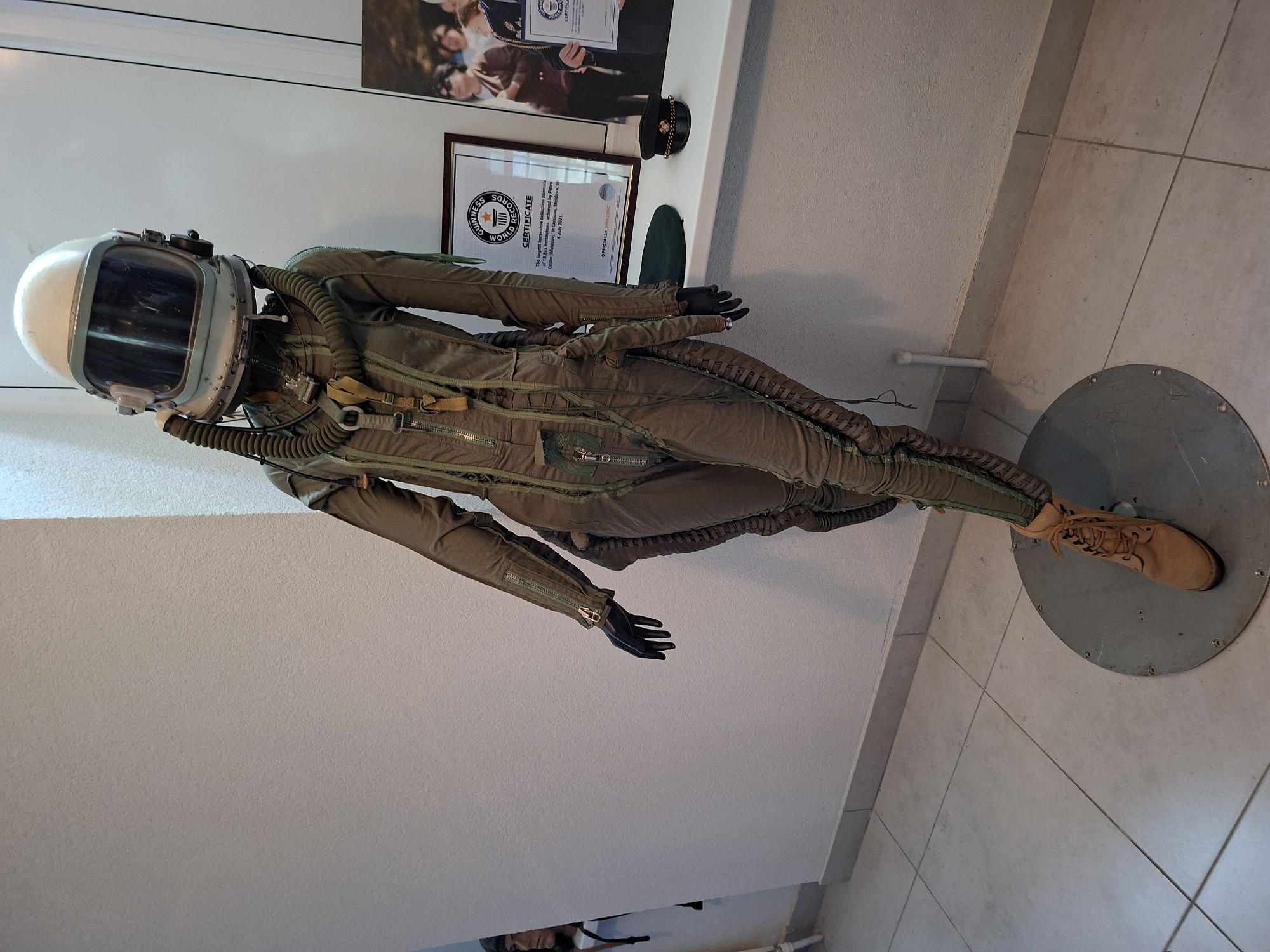 An aviator's flight suit, complete with a helmet and boots, is displayed on a mannequin in what appears to be a museum or exhibition space. The suit is olive green and features numerous zippers, straps, and a ribbed hose extending from the helmet. The mannequin is positioned leaning against a white wall, with its right leg bent and boot visible on a circular metal base. To the right of the mannequin, on a windowsill or shelf, is a framed Guinness World Records certificate, a dark green beret, and a military-style cap. Behind the certificate is a framed photograph of people. Another mannequin, partially visible on the left, wears a dark cap and appears to be looking towards the right. The floor is tiled in a light color. The scene is well-lit, suggesting indoor lighting. The presence of the Guinness World Records certificate indicates a connection to record-breaking achievements, possibly in the context of aviation or a related field. The location is Ialoveni, Moldova, as indicated by the Guinness World Records certificate which states: "The largest horseshoe collection consists of 13,855 horseshoes, achieved by Petru Stefan, Moldova, in Chisinau, Moldova, on 4 July 2021." While the certificate mentions Chisinau and the country Moldova, the specific location in Ialoveni is not directly depicted in the image itself but is provided as contextual information.