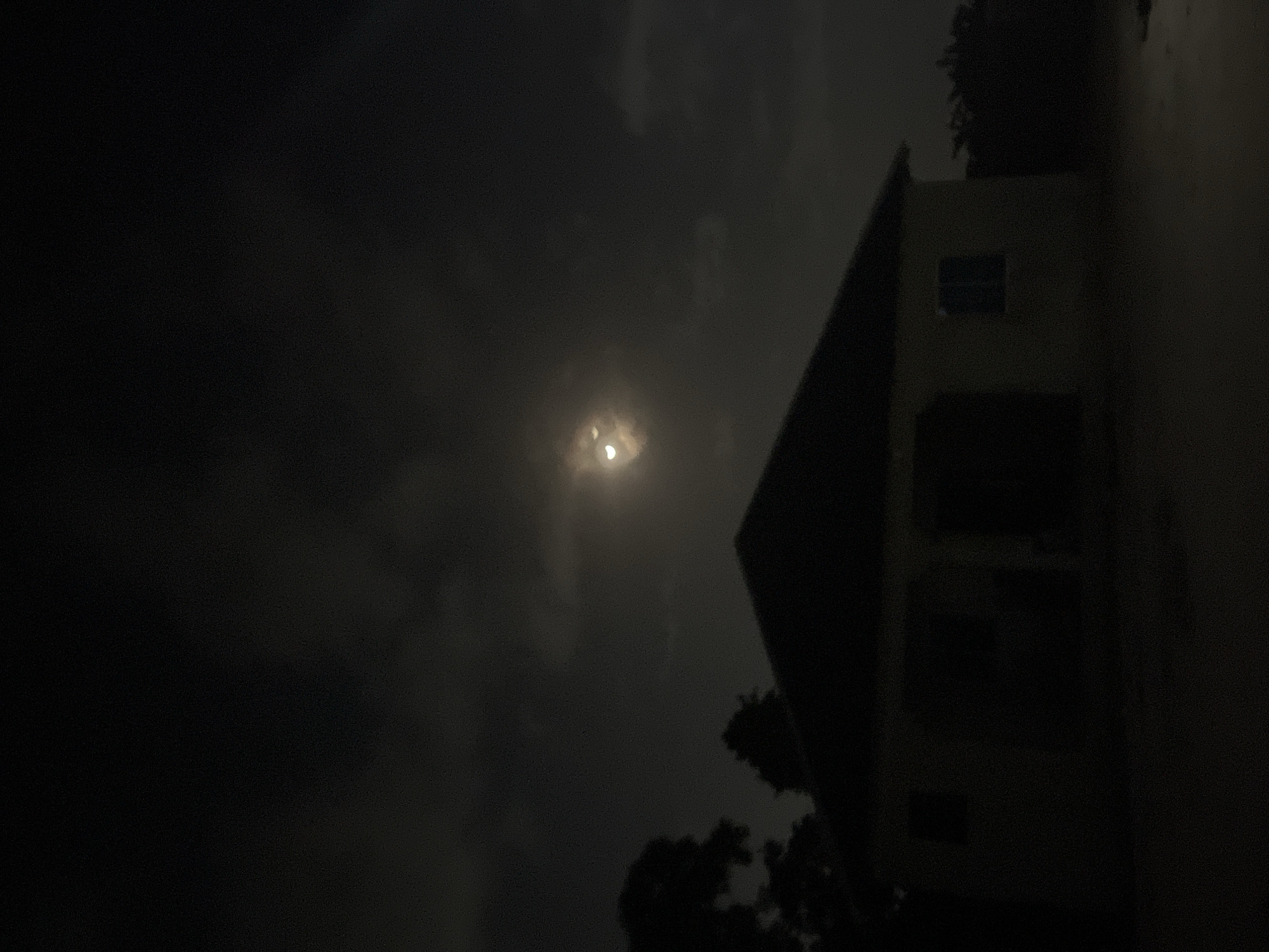 The image shows a night scene in Unknown, Unknown, with a building to the right and a brightly shining moon surrounded by clouds in the sky. The building is two stories high, with visible windows and a roofline. Trees are partially visible in the foreground and behind the building. The overall lighting is dim, with the moon being the primary light source.