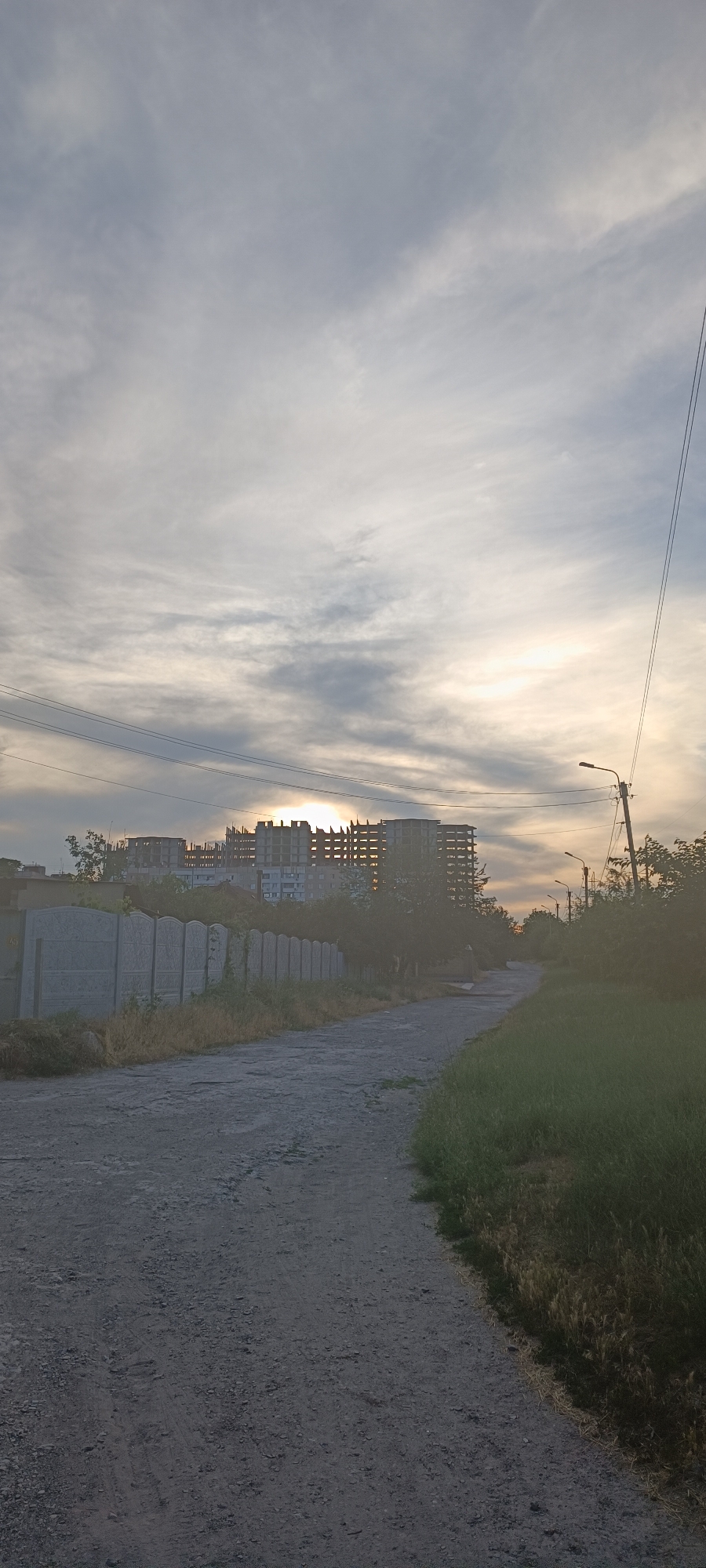 The image depicts a scene at dusk in Zaporizhzhia, Ukraine, with the sun setting behind unfinished buildings under a cloudy sky. The foreground features a gravel road winding through an area with a concrete fence on the left and overgrown grass on the right. In the background, several multi-story buildings are under construction, with their skeletal structures visible against the sky. Electrical wires and a street lamp are also present, suggesting an urban or semi-urban environment. The time of day is evident from the low angle of the sun and the soft, diffused light filtering through the clouds, casting a warm glow on the scene. The overall mood is one of quiet anticipation or transition, possibly reflecting the ongoing development of the area. No people are visible in the image. There is no visible text.