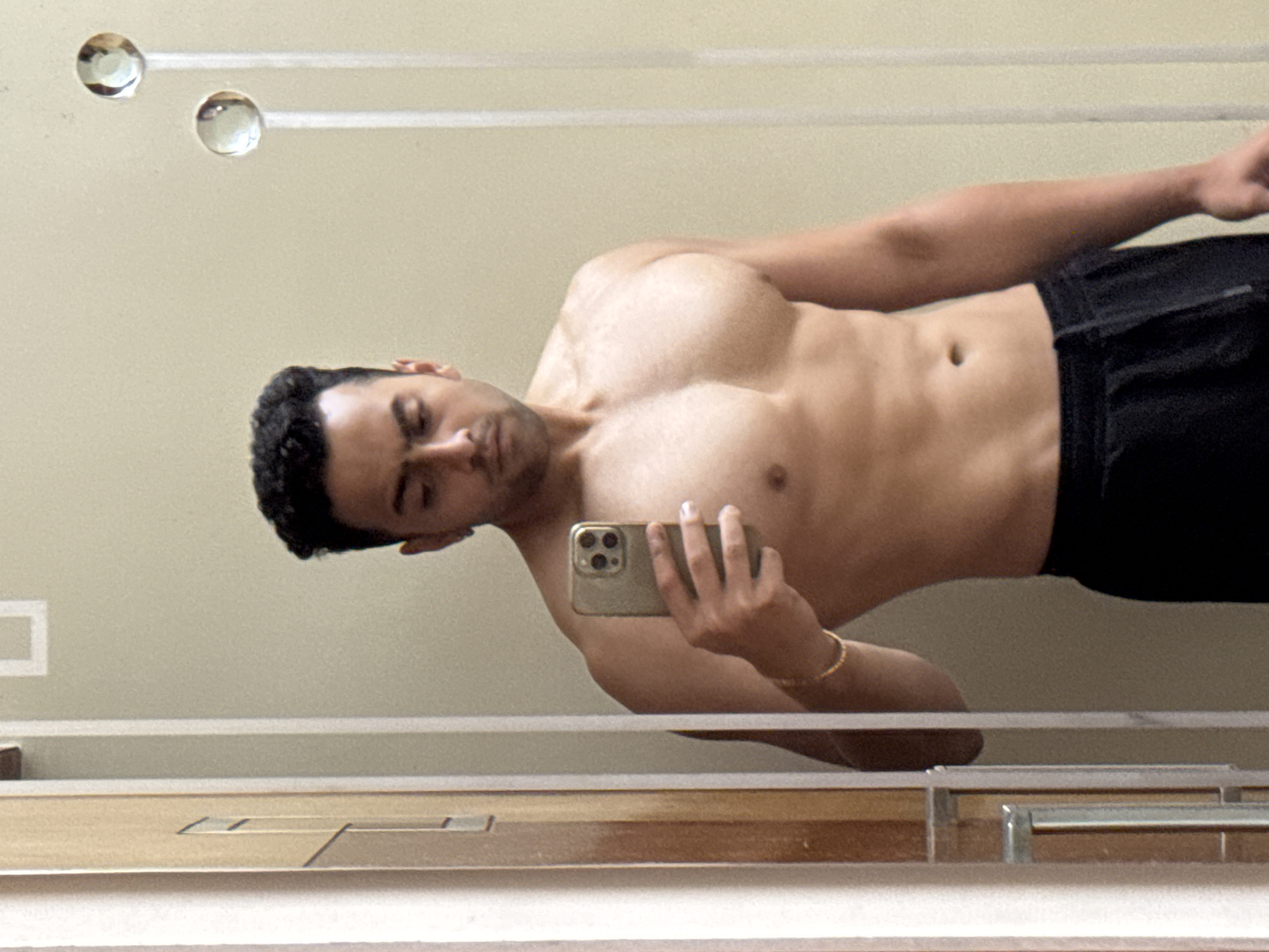 A man is taking a selfie in a bathroom mirror. He is shirtless and wearing dark shorts. The background is a plain, light-colored wall with decorative glass knobs on a horizontal bar. The bathroom vanity has a wood-grain finish. The location in Kolkata, India is not discernible from the image.