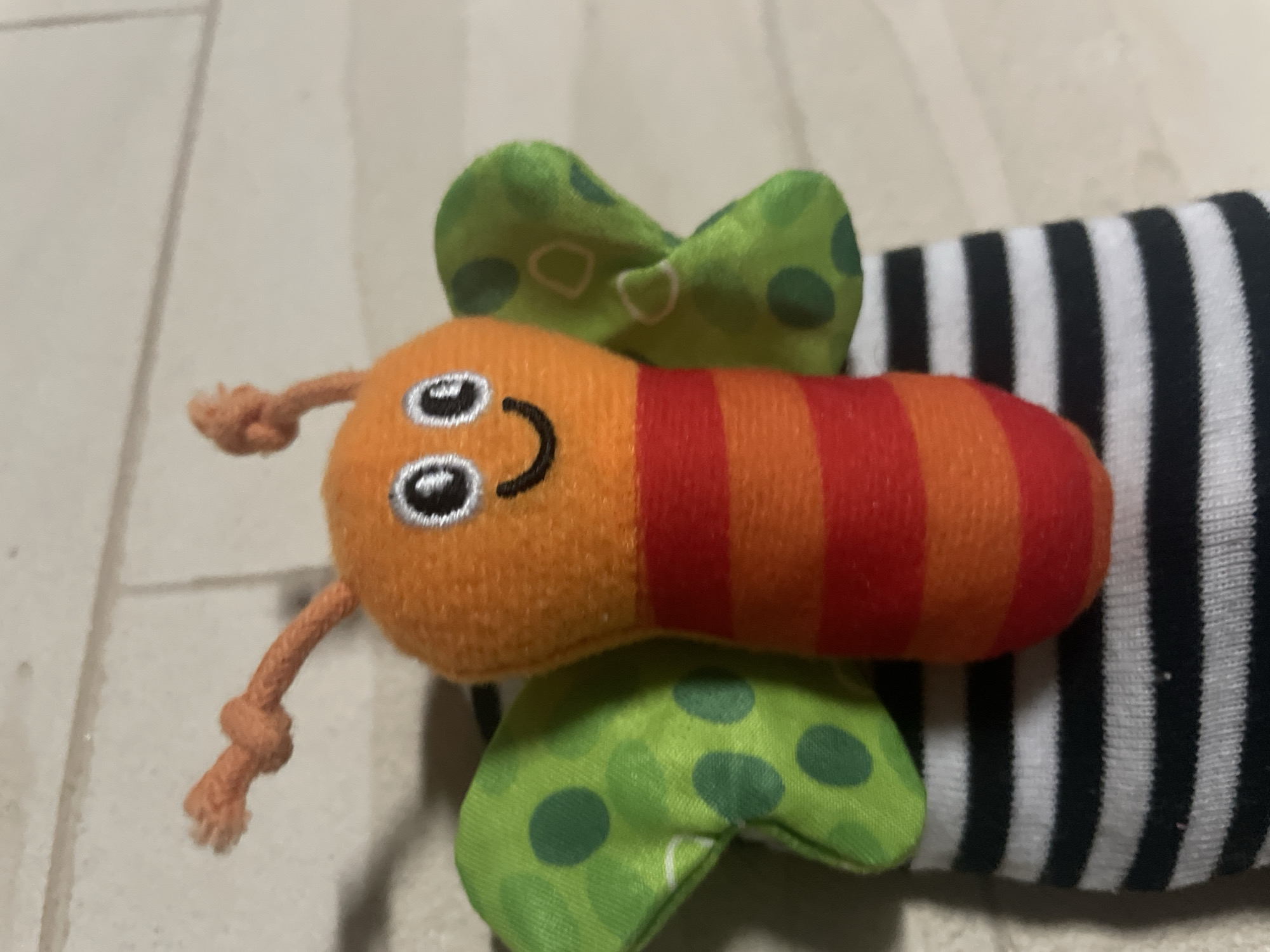 A close-up photograph depicts a plush toy caterpillar. The caterpillar's head is orange with embroidered eyes and a smiling mouth. The body is segmented with alternating orange and red stripes. Green polka-dotted wings extend from the caterpillar's body. To the right of the caterpillar is a black and white striped fabric. The background is a light-colored, possibly wooden, surface. The location is unknown, Novi Sad, Serbia is not inferable from the image.
