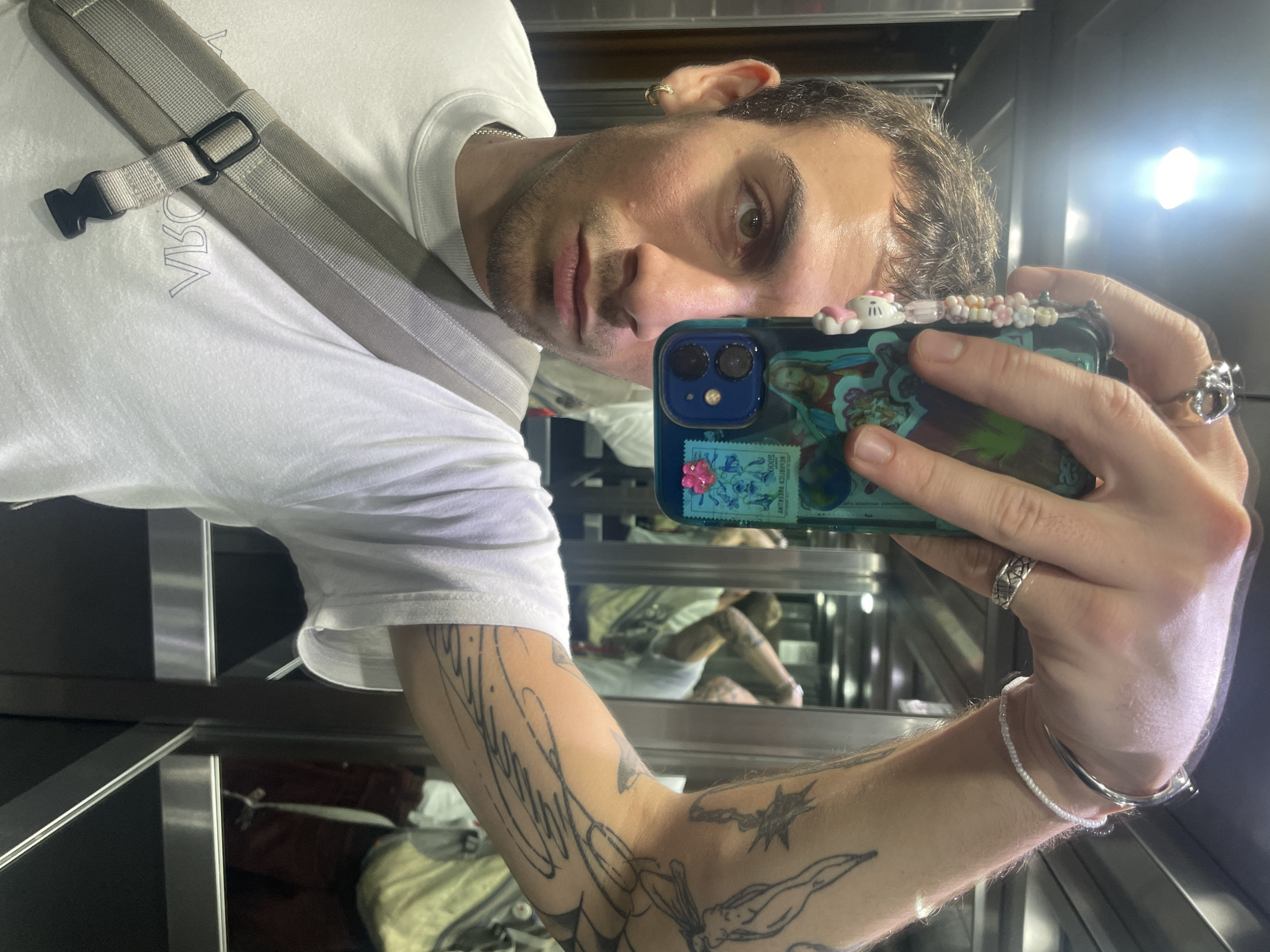 A light-skinned man with short brown hair and light brown eyes takes a selfie in a mirrored elevator. He is wearing a white t-shirt with a gray shoulder strap across his chest. He holds a phone with a blue case decorated with stickers and beads. Tattoos are visible on his arm. The reflection in the elevator mirrors the man and other objects within the elevator space. The location is Buenos Aires, Argentina.