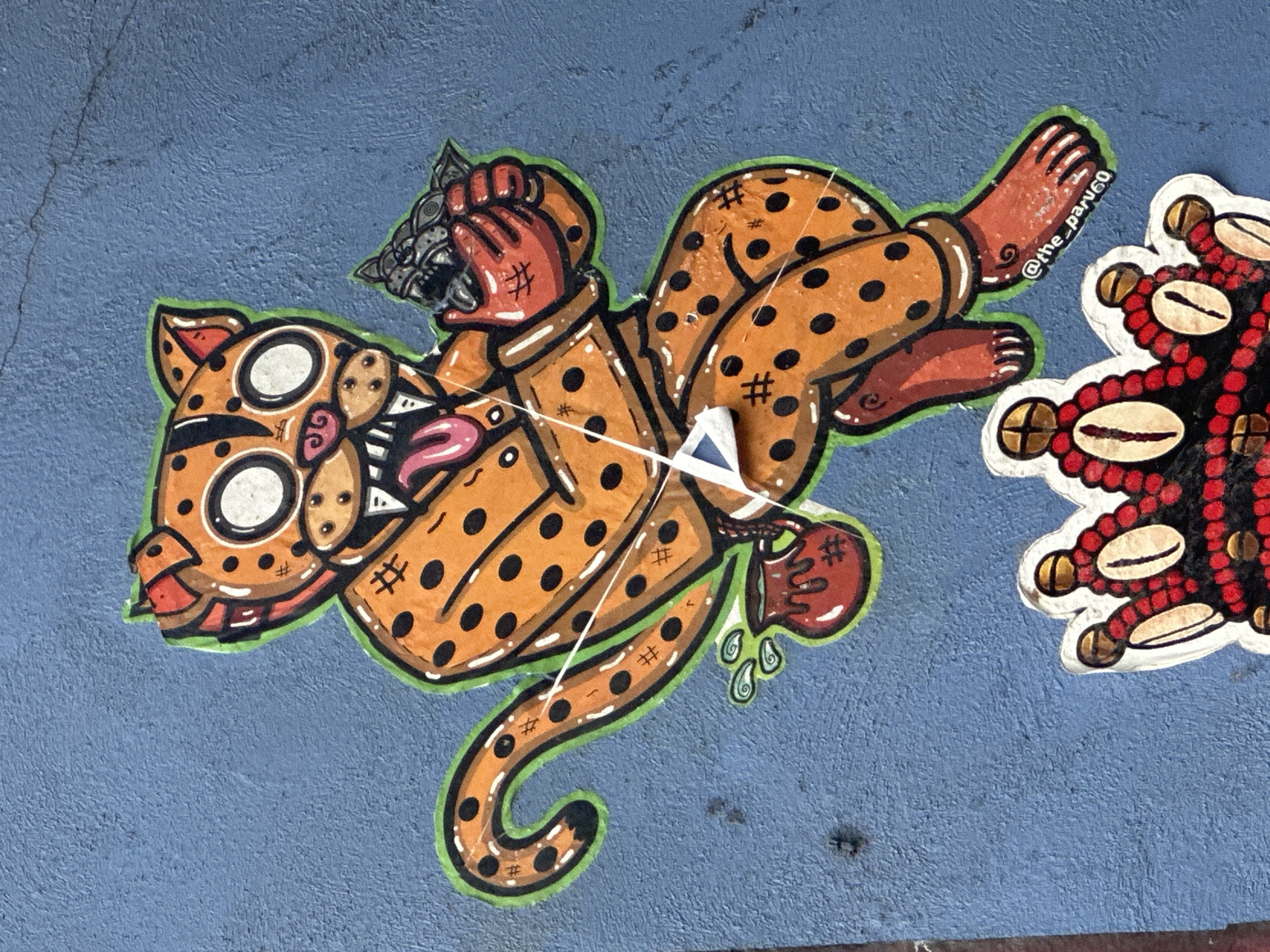 A close-up shot reveals a piece of street art in Mexico City, Mexico. The artwork features an anthropomorphic orange jaguar with black spots and green outline lying on a blue concrete wall. The jaguar has cartoon-like eyes, a pink tongue, and is depicted wearing a dark jacket and clutching a small, possibly stone, jaguar head in its hand. One of its paws is adorned with hash symbols. A small, red pot is attached to the jaguar's waist, with some drops falling from it. A string of white thread hangs from the triangle attached to the jacket. To the right of the jaguar is a beaded object in red, black, and white. The artist's tag "@the_paru60" is written near the right paw of the jaguar. The surface of the concrete wall has a textured appearance.