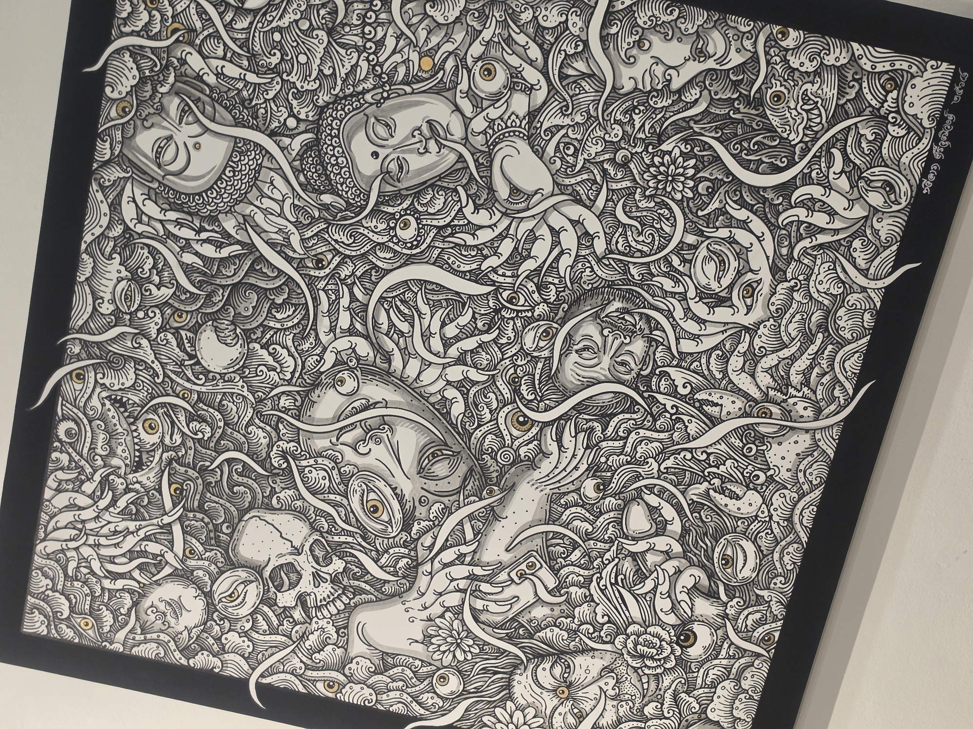 The image shows a framed, detailed black and white illustration. The illustration features multiple stylized human faces, skulls, and eyes interspersed within swirling, organic patterns resembling waves, tendrils, and floral elements. The faces have a serene or contemplative expression. The eyes scattered throughout the composition are detailed with defined irises. The image is framed in black. The location is not specified.