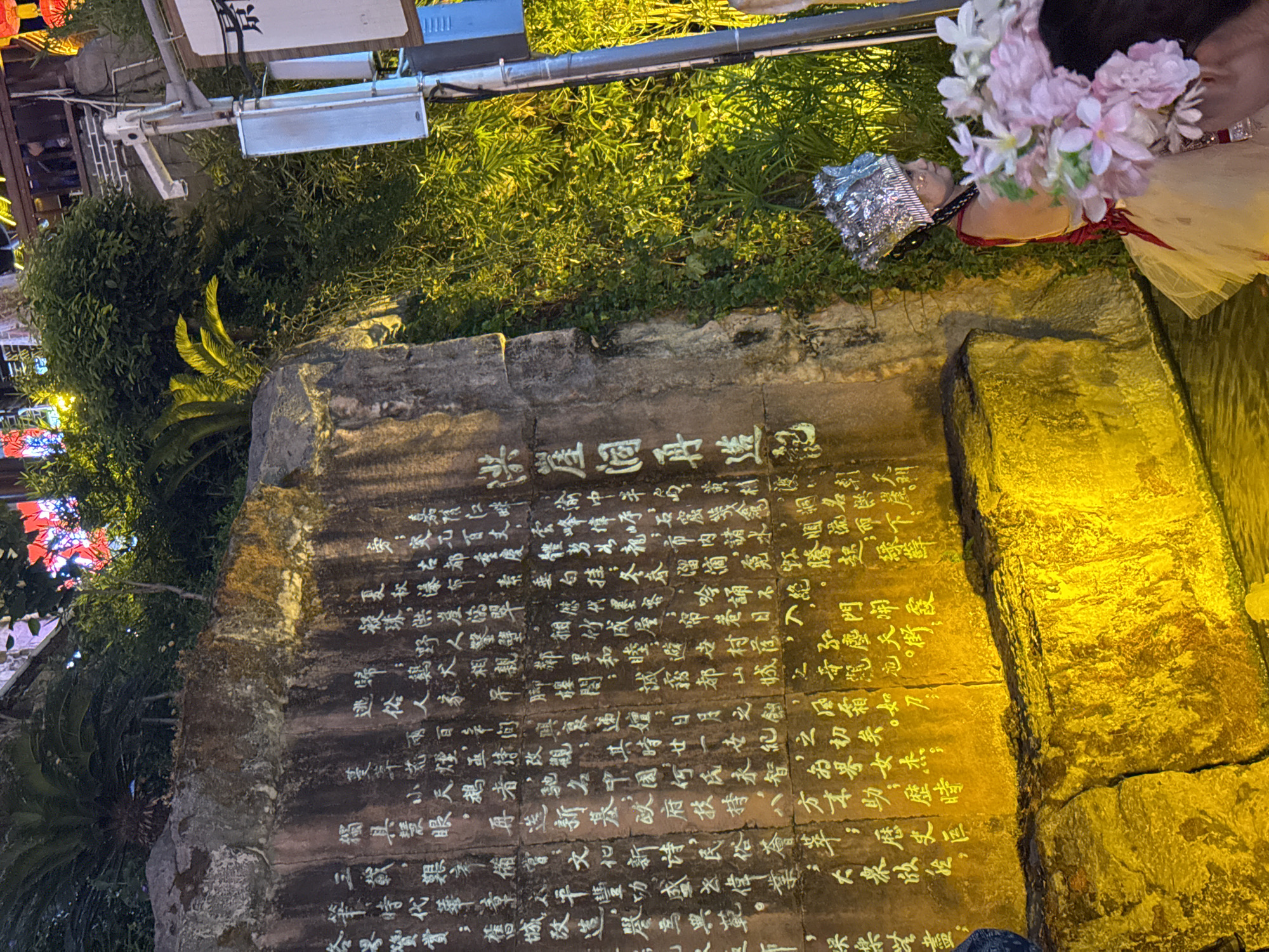 The image shows a large rock face inscribed with Chinese characters, illuminated by yellow lights. The rock is situated outdoors, adjacent to lush green foliage and what appears to be water at its base. In the foreground, a person wearing an ornate silver headdress and a light-colored dress is visible on the right side of the frame, holding a bouquet of pink and white flowers. Behind them, the rock face is extensively covered with white calligraphy. The scene suggests an outdoor cultural or historical site, possibly in the Yuzhong District of China, given the presence of traditional Chinese script and the architectural style visible in the background, which includes lighting fixtures and buildings. The lighting creates a dramatic effect on the rock and vegetation.