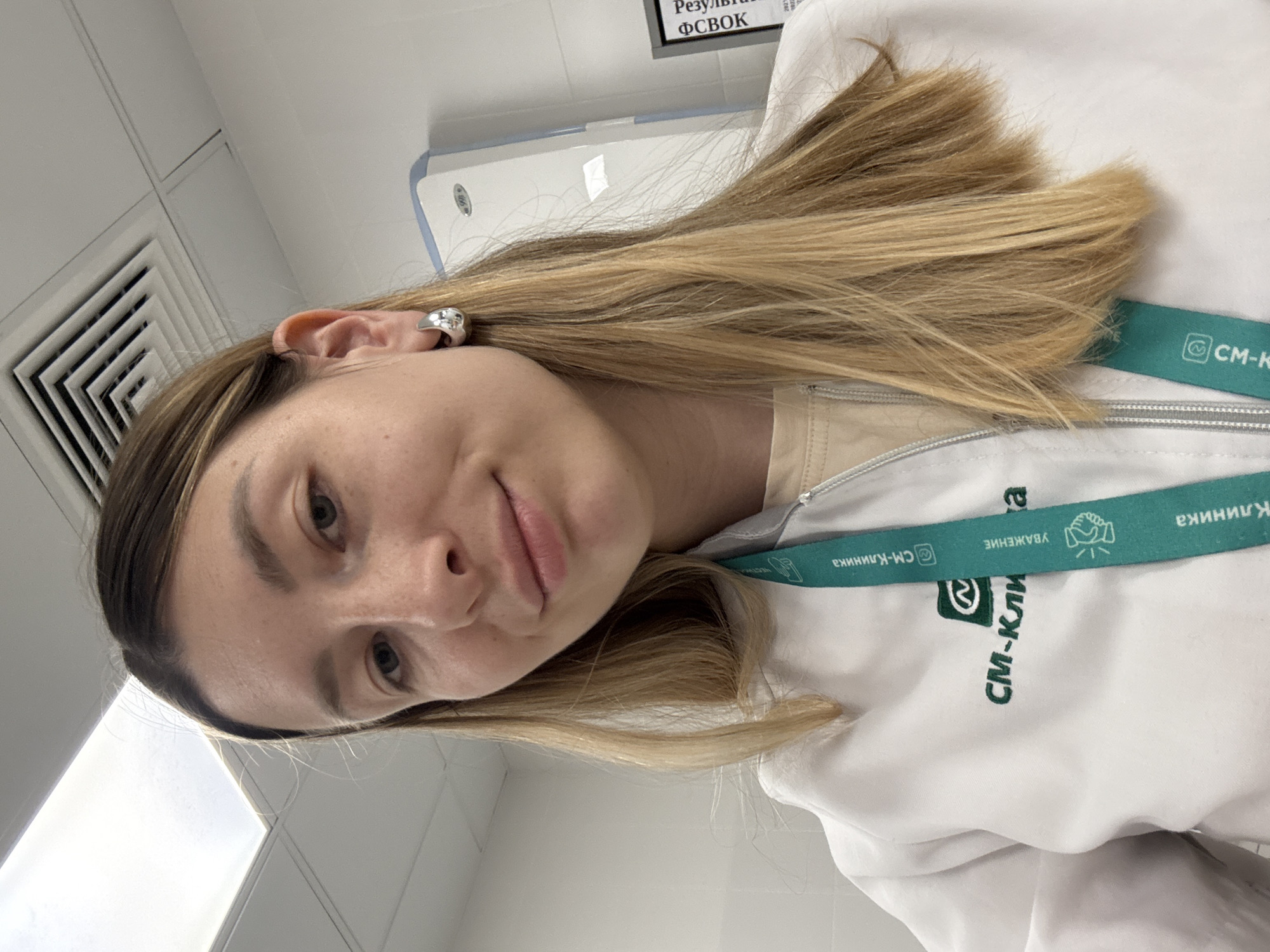 A young Caucasian woman with long blonde hair and fair skin is pictured in a close-up shot. She is wearing a white lab coat with a green lanyard around her neck. The lanyard has the logo "СМ-Клиника" and other text. She is also wearing a silver hoop earring on her left ear. In the background, a white wall and a ceiling vent are visible. A document with the word "Результат" is partially visible on the right side of the background. The lighting suggests an indoor environment, possibly a clinic or hospital setting in Moscow, Russia.