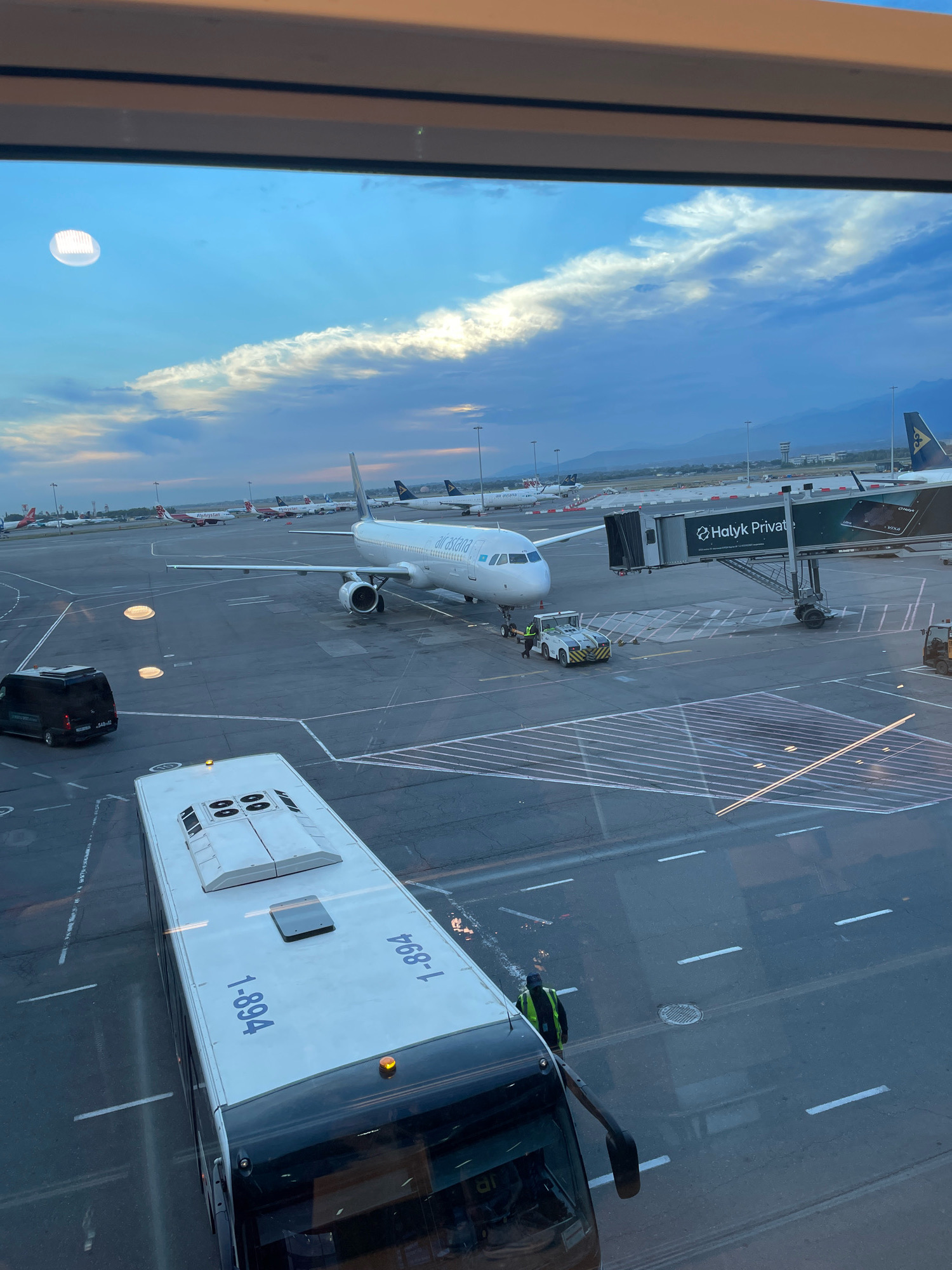 The image depicts an airport tarmac scene in Almaty, Kazakhstan, during either dawn or dusk, as indicated by the soft, angled light and the presence of both bright and muted sky colors. In the foreground, a white passenger bus with the number "1-894" visible on its roof is parked. To the left, a black van is also stationary.
Dominating the mid-ground is a white passenger airplane with "Air Astana" branding on its fuselage. The aircraft is connected to a jet bridge, labeled "Halyk Privat," which leads to a terminal building. Several airport service vehicles, including a small tug, are positioned near the airplane. In the background, numerous other aircraft are parked on the tarmac, and in the far distance, a control tower and mountainous terrain are visible. The sky features wispy clouds illuminated by the setting or rising sun, casting long shadows across the asphalt. The overall scene captures the operational activity of an airport at a transitional time of day.