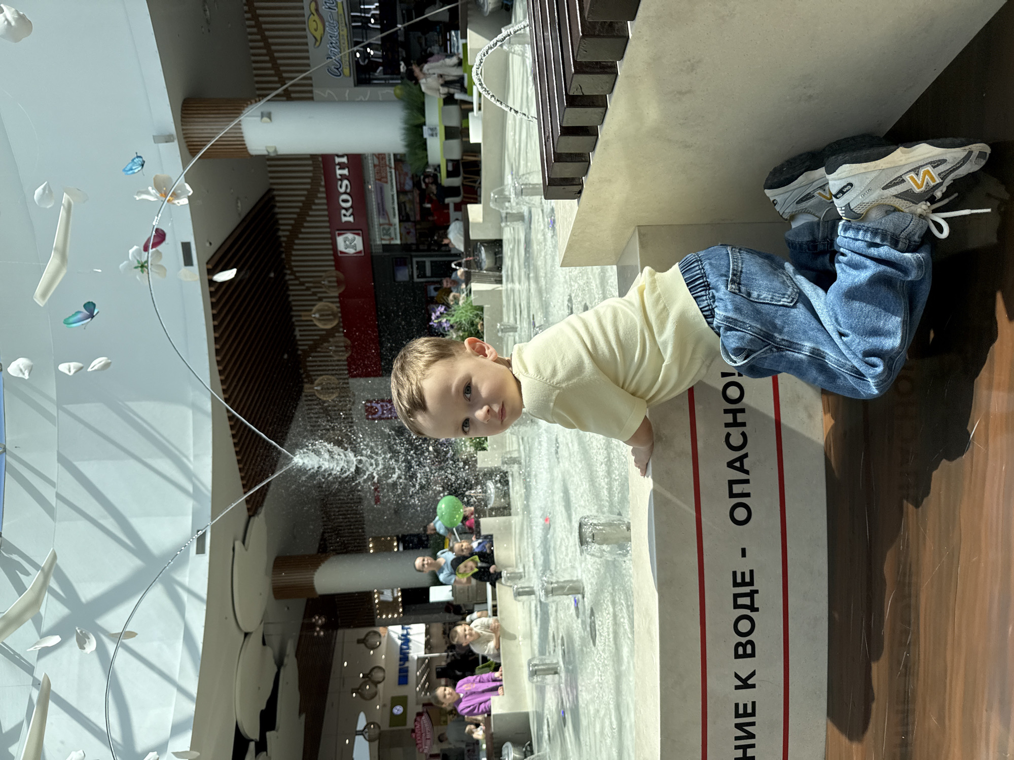 A young child is kneeling at the edge of an indoor fountain in Vladivostok, Russia, looking towards the camera. The child is wearing a light yellow shirt and blue jeans, and has on gray and white sneakers with a yellow "N" logo. The fountain has multiple small water jets and is located inside a shopping mall. Visible behind the fountain are various stores and people. A sign in Russian is next to the child.