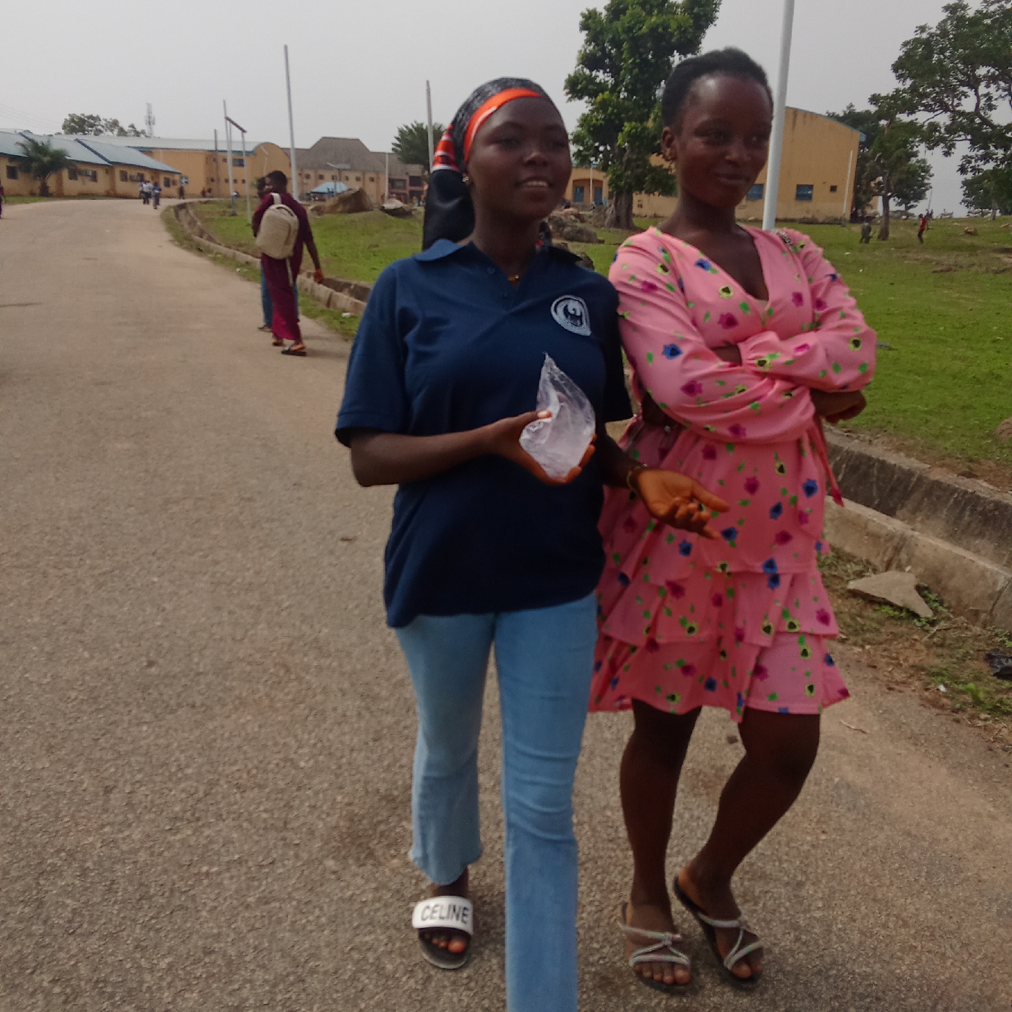 Two young women walk on a paved road in what appears to be Akwanga, Nigeria. The woman on the left wears a dark blue short-sleeved shirt with a logo, light blue jeans, and "CELINE" branded sandals. She has an orange headband and holds a clear plastic bag in her left hand. The woman on the right wears a pink floral dress with ruffles and sandals. Her arms are crossed. In the background, other people walk along the road towards yellow buildings and green grassy areas with trees. Poles stand near the buildings. The sky is overcast. A concrete curb runs along the edge of the road near the woman on the right.