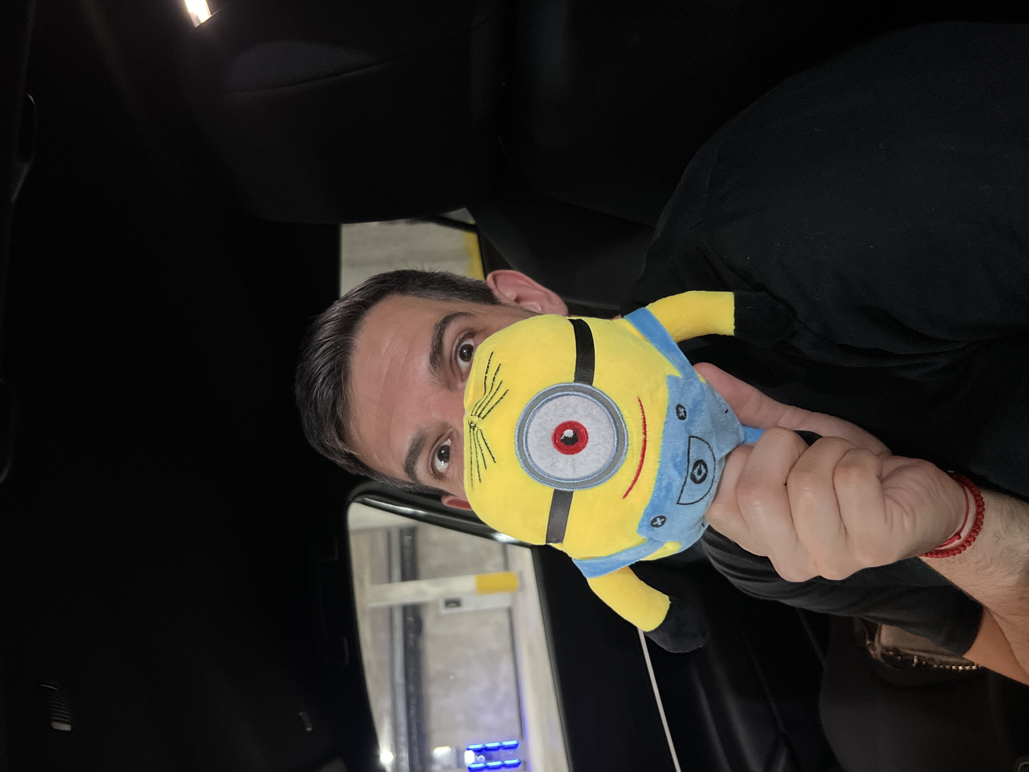 A man is sitting in the driver's seat of a car, holding a plush minion toy in front of his face, peeking over it with wide eyes. He is wearing a black t-shirt and a red bracelet on his left wrist. The car's interior is dark, with the ceiling and headrest visible. Outside the car, a parking garage with concrete pillars and some blue lighting can be seen.