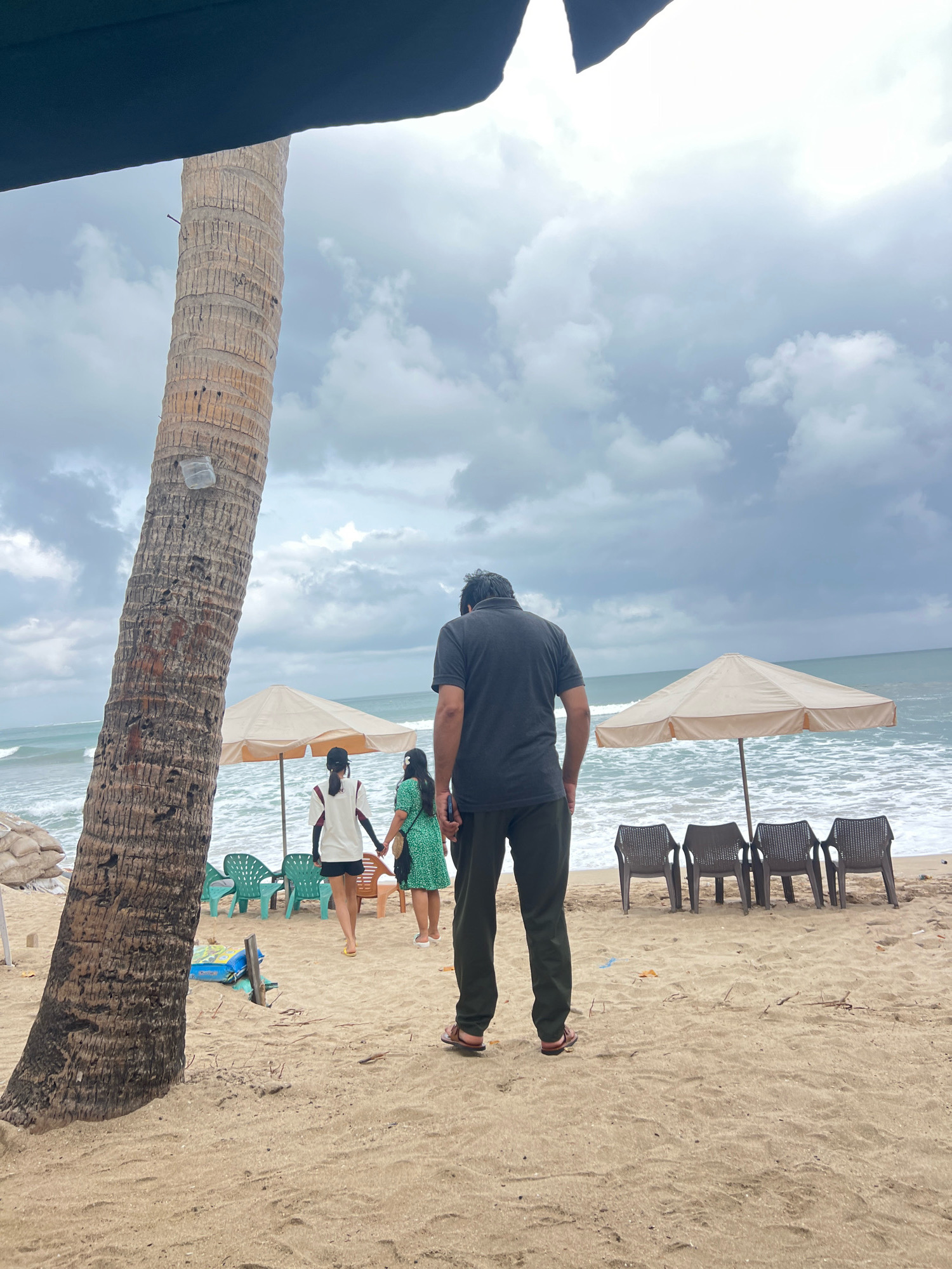 The image shows a beach scene in Kuta, Indonesia, with a man standing in the foreground looking out at the ocean. Behind him, two women are walking hand-in-hand on the sand. There are beach umbrellas and chairs set up on the sand, and the ocean waves are visible in the background. The sky is cloudy. A large palm tree is to the left of the frame, and some sandbags are piled up in the distance.