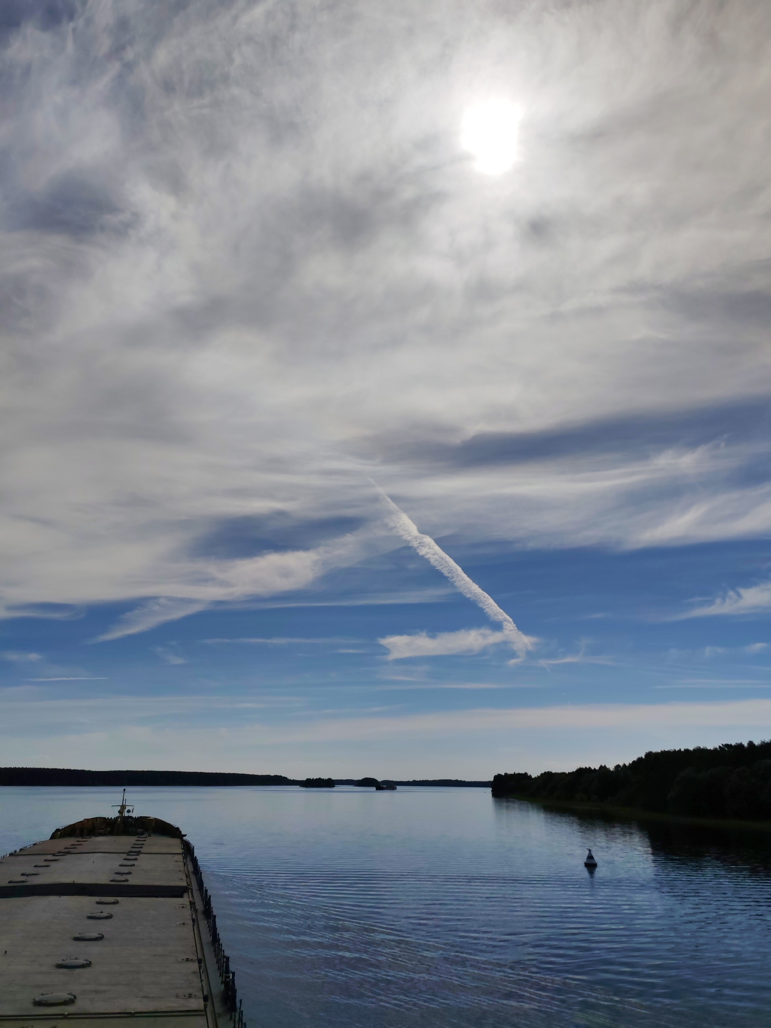 The image depicts a broad daylight scene from the forward deck of a large vessel, likely a cargo ship or barge, traversing a wide body of calm water in the vicinity of Domazhino, Russia. The sky is largely overcast with a mix of stratiform and cirrocumulus clouds, through which the sun emits a bright, intense glare in the upper right quadrant. A prominent, white aircraft contrail diagonally bisects the mid-sky, forming a distinct linear cloud formation. The horizon features distant, dark green treelines along both banks of the waterway, with hints of smaller islands in the distance to the left. The water's surface reflects the sky's blue and grey hues, showing subtle ripples. A solitary white buoy or navigational marker is visible in the water on the right side, nearer to the forested bank. The foredeck of the vessel, which occupies the lower left frame, is a flat, grey surface with evenly spaced rectangular hatches or covers and a small mast-like structure at its very front. No individuals are visible on the vessel or in the scene.
