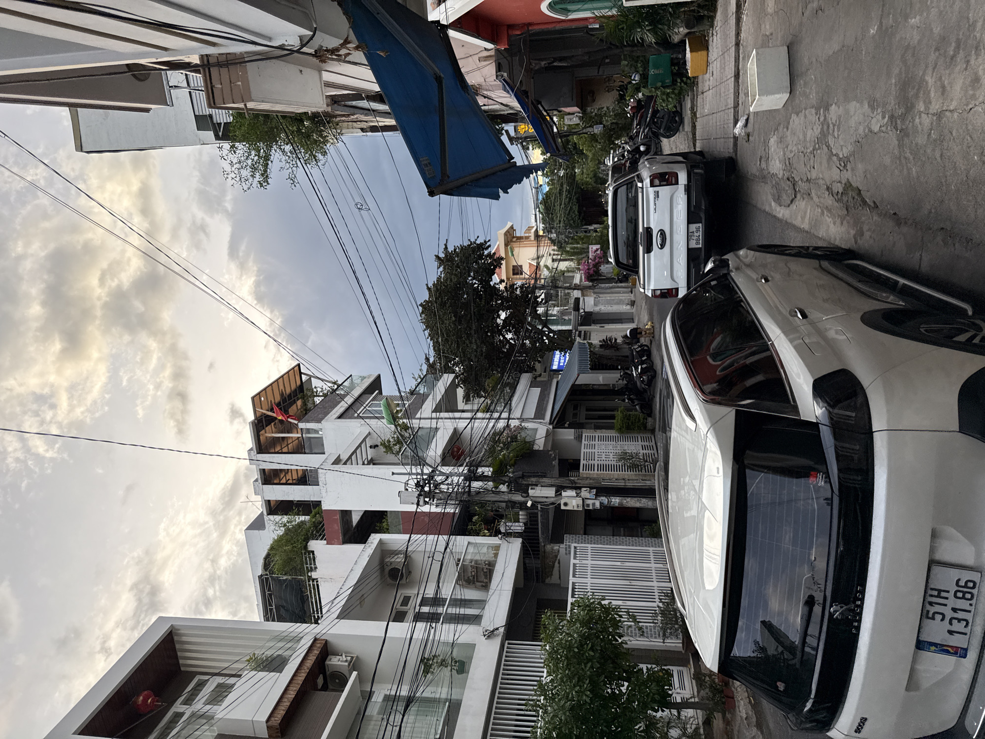 The image shows a narrow street in Nha Trang, Vietnam, lined with multi-story buildings. The sky is overcast with dramatic clouds. In the foreground, a white Peugeot SUV is parked, partially obscuring the view of a white Ford Ranger pickup truck behind it. The Ford Ranger has a license plate reading "79A 884 96". A white concrete planter box is visible on the sidewalk to the right of the Ford Ranger. The buildings on both sides of the street have balconies, air conditioning units, and a dense network of overhead electrical wires. Some motorcycles are parked along the sidewalks, and a few are also visible further down the street. The architecture of the buildings appears to be a mix of modern and traditional styles. A red lantern hangs from the exterior of a building on the left. The overall impression is that of a typical urban street scene in Vietnam.