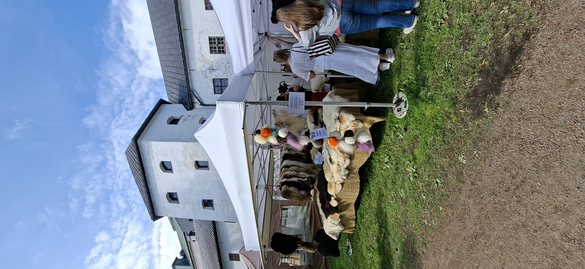 An outdoor market stall displays a variety of animal pelts and colorful pom-poms under a white canopy. The stall is set up on a grassy area, with a gravel path in the foreground. In the background, a historic white building with a dark roof and arched windows is visible. Two women are browsing the stall, one in a white dress and the other in a grey shirt and blue jeans. The location is confirmed to be Turku, Finland, likely at the Turku Castle market, a popular historical reenactment and craft fair event.