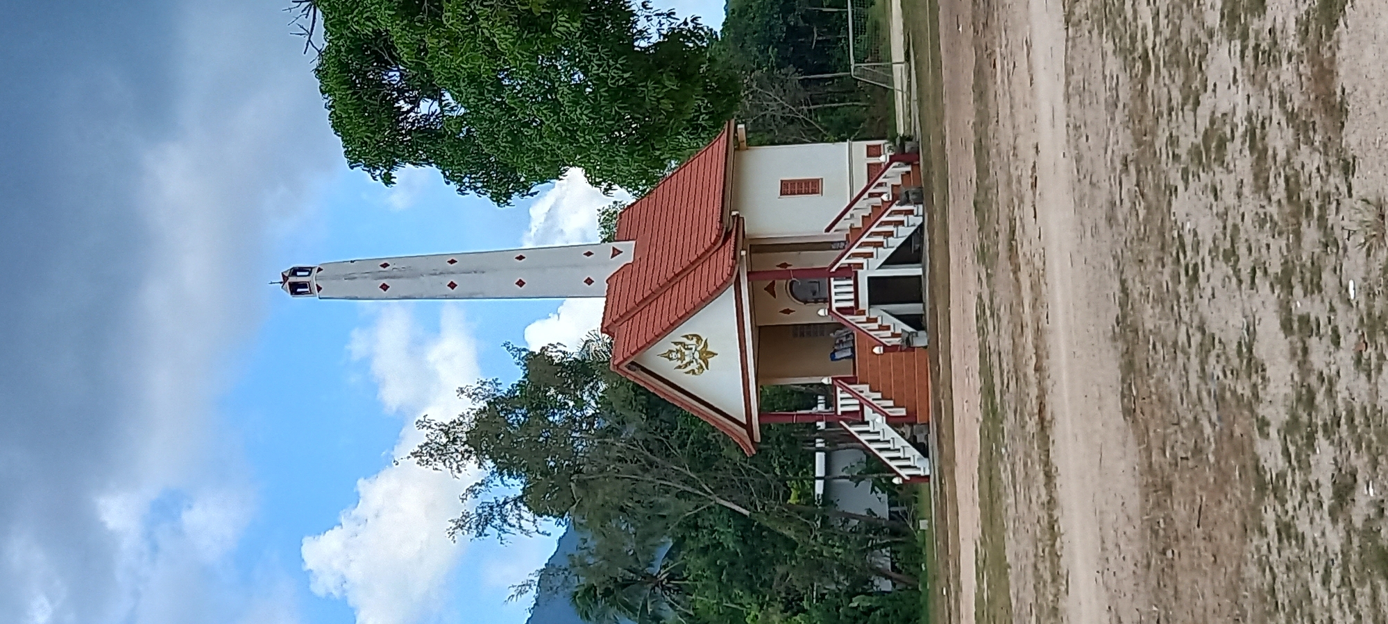The image displays a serene daytime scene in what appears to be Baan Chaloklum, Thailand, featuring traditional architecture amidst natural surroundings.
In the mid-ground, a two-story building with a distinctive red-tiled roof and white walls stands prominently on the right. Its front gable is adorned with an intricate golden symbol, likely a Buddhist motif. Two symmetrical sets of white staircases with red handrails lead up to the second-story entrance. The ground floor of the building is recessed, and windows are visible on the upper level.
To the left of this building, a tall, slender white tower rises vertically, featuring regularly spaced red diamond-shaped markings along its length. The top of the tower has dark openings or decorative elements. Given the context, this structure could be a crematorium chimney or a stupa/chedi associated with a local temple.
The scene is framed by abundant green trees, with a large, leafy tree occupying the upper left quadrant and other foliage surrounding the buildings. The sky is a vibrant blue, dotted with scattered white clouds, indicating clear weather. The foreground and the area to the right of the building consist of a flat, earthy expanse with patches of green grass, resembling an open field. In the far background, a mountain or large hill is visible beneath the sky. A faint soccer goalpost frame can be seen in the distant background on the right, suggesting a recreational area nearby.
No people are visible in the image, and there is no discernible text on any of the structures. The overall atmosphere conveys a sense of tranquility and local cultural heritage.