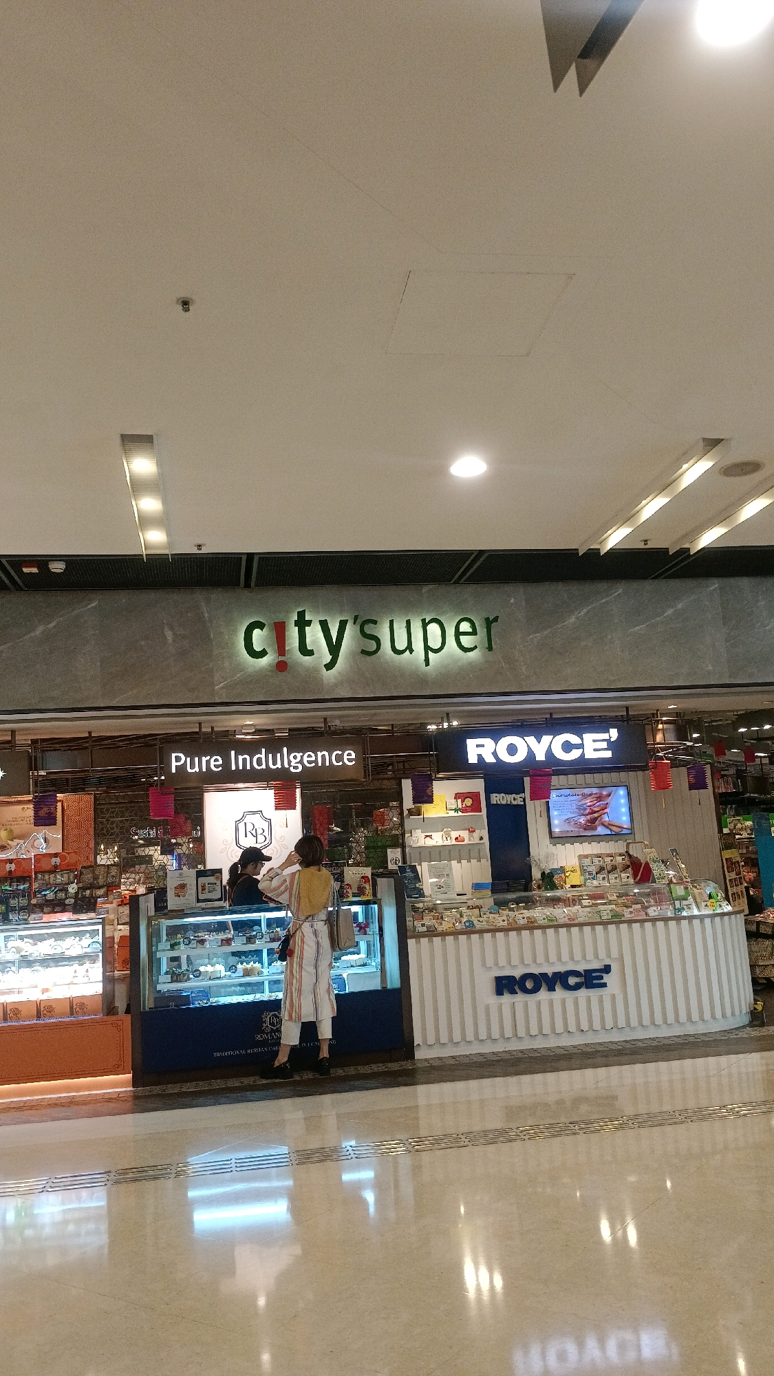The image shows the interior of a City'super store in Hong Kong, China. In the center, a woman wearing a striped outfit and a headscarf stands behind a display case filled with cakes and pastries. She appears to be serving a customer. To the right of the cake display is a Royce' chocolate shop with a prominent blue and white sign. The store is well-lit, with a polished floor reflecting the lights and displays. The overall environment suggests a modern and upscale retail space.