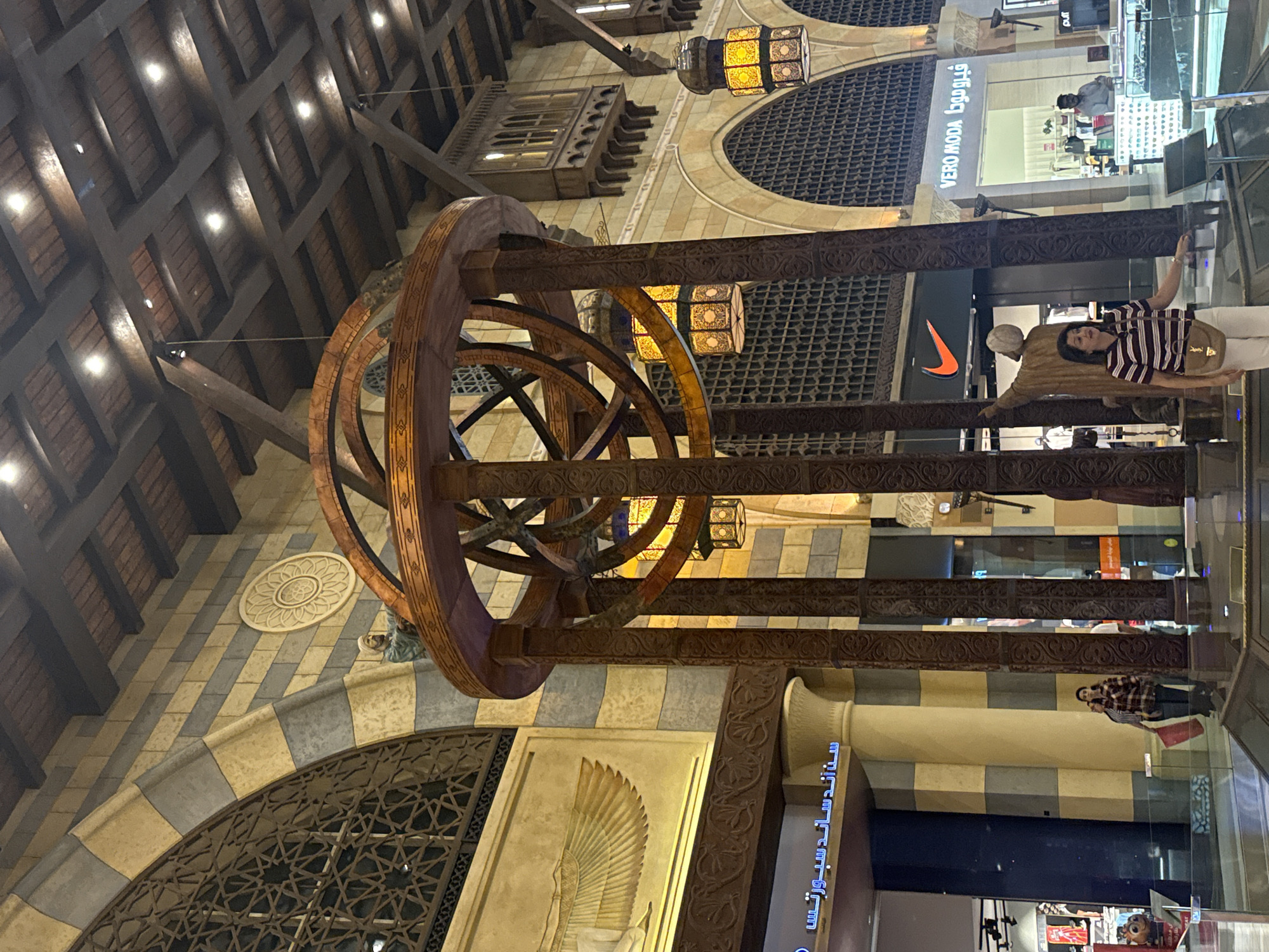 In Dubai, United Arab Emirates, an ornate, large-scale astrolabe-like sculpture is prominently displayed in the center of a grand, indoor marketplace. The sculpture features dark wood, intricately carved pillars supporting concentric rings, and decorative lanterns. The surrounding architecture reflects a traditional Middle Eastern aesthetic with arched doorways, patterned stonework in beige and blue, and a wooden beamed ceiling with modern lighting.
To the left of the sculpture, a large wall mural depicts a golden winged figure, likely an Egyptian motif, and a decorative circular relief. Adjacent to this, a storefront displays Arabic text and a golden winged emblem. Further to the left, a blue and black storefront is visible, with some signage in Arabic.
To the right of the central sculpture, a statue of a person in traditional attire stands near a storefront with the Nike logo. Further right, a storefront labeled "VERO MODA" in Arabic and English is visible, along with other retail displays.
Several individuals are present in the scene. In the foreground, a woman wearing a striped polo shirt and white pants is standing near the sculpture, looking to her left and holding a tan handbag. Behind her, another person is partially visible. Further in the background, other shoppers are seen walking and browsing in the retail areas. The overall environment suggests a busy, upscale shopping destination.
