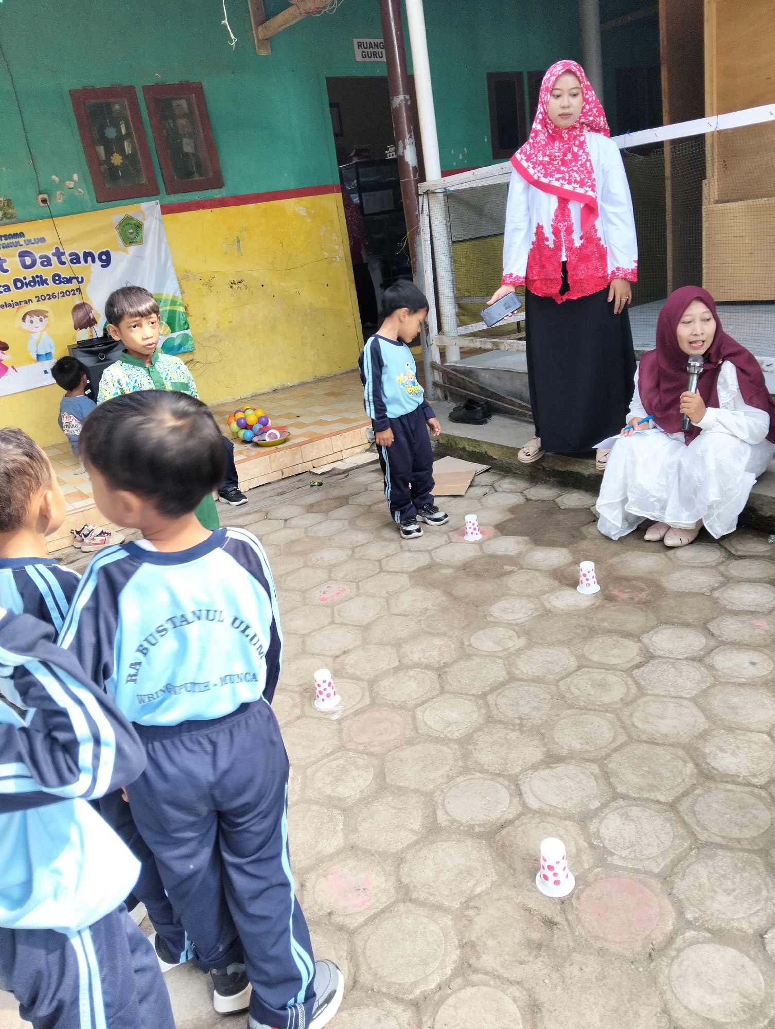 In Desa Wringin Putih, Indonesia, an outdoor school event is underway. Two women, one wearing a pink and white headscarf and the other a maroon headscarf, are interacting with a group of young children. The woman in the pink and white headscarf holds a smartphone and appears to be addressing the group, while the woman in the maroon headscarf is seated and holding a microphone and pen, looking towards the children. Several children, dressed in school uniforms, are engaged in an activity on the ground. The ground is paved with hexagonal concrete blocks. Scattered on the ground are small, decorated paper cups, suggesting a game is in progress. In the background, a yellow banner with Indonesian text and cartoon illustrations is visible, along with green and teal painted walls. The overall scene depicts a lively educational gathering in a schoolyard.