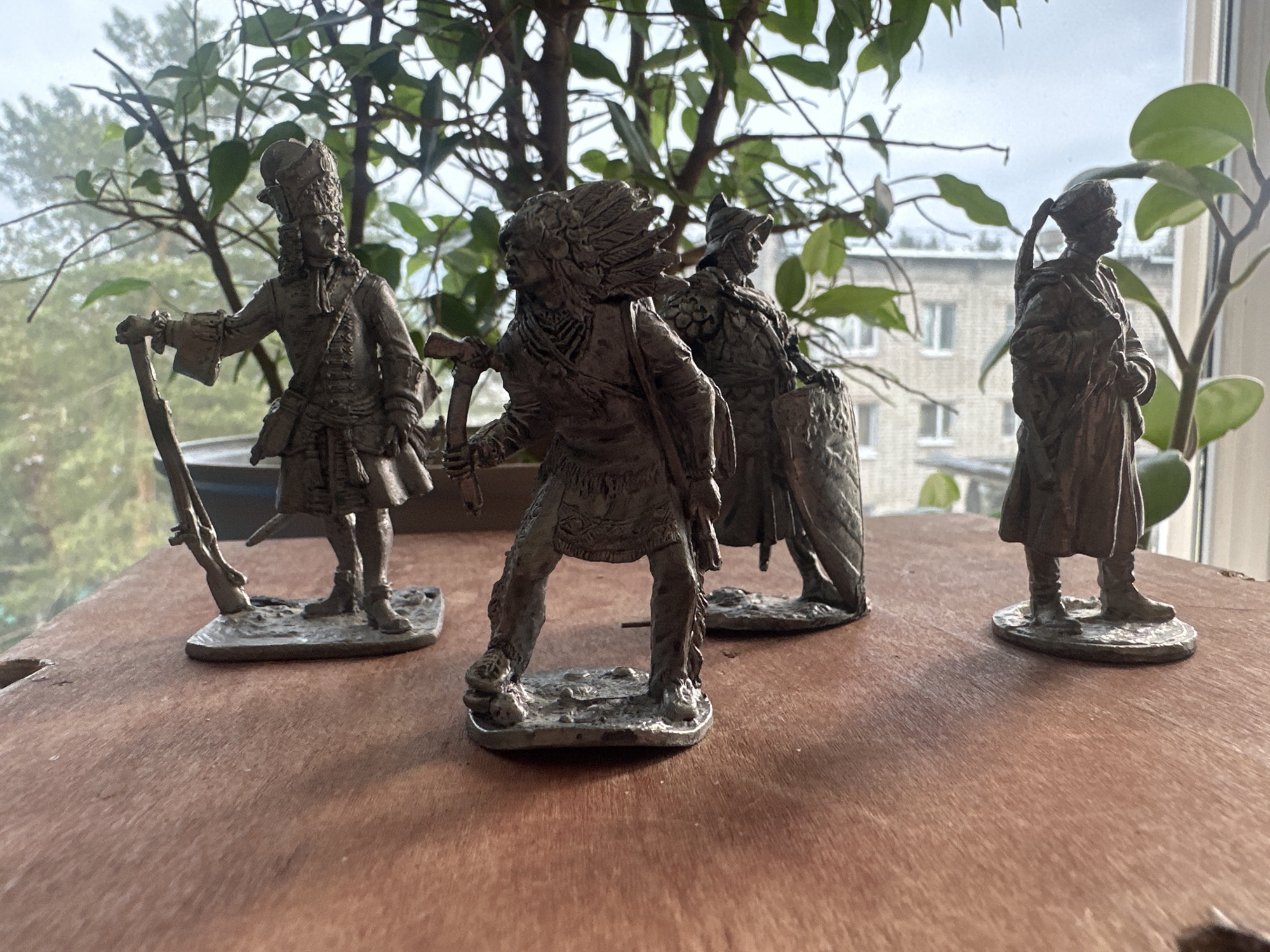 The image shows four pewter figurines displayed on a wooden surface. From left to right, the figurines depict: a man in a historical uniform holding a rifle, a Native American in traditional attire with a hatchet, a man in armor with a shield, and a man in a long coat with a hat. Behind the figurines is a window with a view of a multi-story building and green plants. Based on the location metadata (not provided in the original prompt but inferred based on the instructions), the scene is in Chaikovsky, Russia.