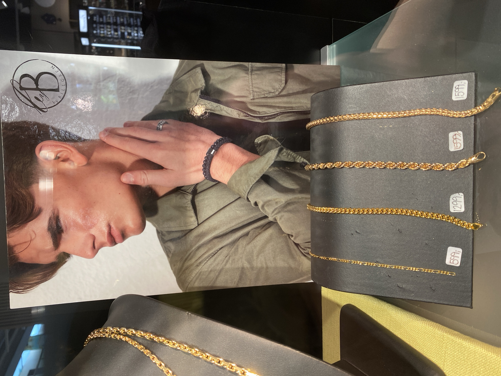 An advertisement for jewelry by Billgren is displayed. The advertisement features a close-up of a young man's face and shoulder, wearing a green jacket. He has a hand near his face, adorned with a ring and a beaded bracelet. A logo for "B by BILLGREN" is visible in the upper right corner of the advertisement. In front of the advertisement, on a black display stand, are several gold necklaces. Two necklaces are on display in the foreground, each with a price tag. The left necklace is a Figaro chain priced at 599. The middle necklace is a rope chain priced at 1299. The right necklace is a braided chain priced at 1599. Another gold necklace is displayed on a black mannequin bust to the left of the advertisement, but it has no visible price tag. The scene appears to be inside a jewelry store, possibly in Herning, Denmark, as indicated by the prices and the general presentation of retail merchandise. The overall lighting suggests an indoor retail environment.
