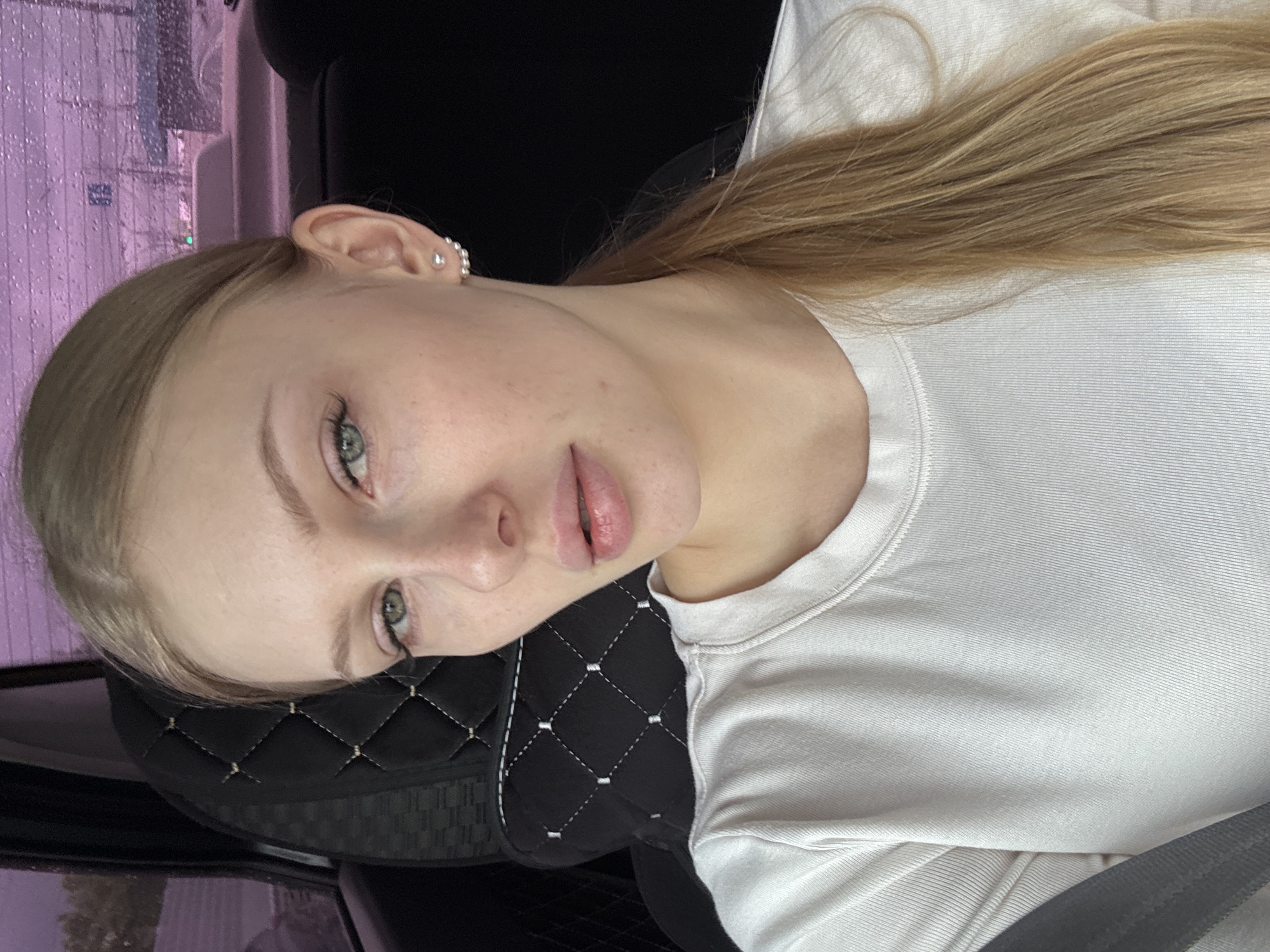 A light-skinned young woman with blonde hair is in a car. She wears a white shirt and is seated in a black car seat with quilted stitching. A row of buildings and cars is visible outside the car window, with raindrops on the glass. This is in Perm, Russia.