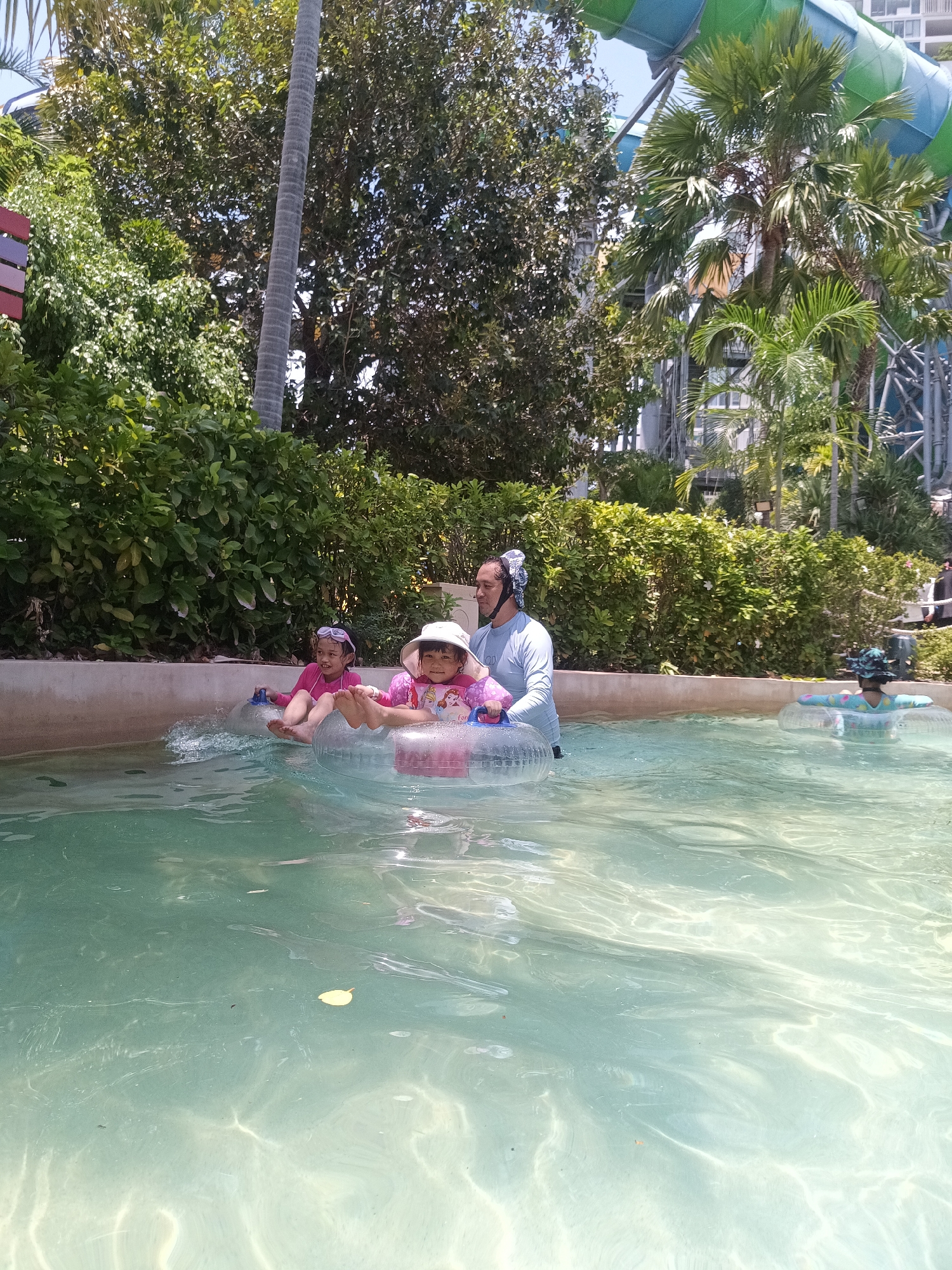 The image shows a family enjoying a day at a water park. Two young girls, wearing swimsuits and hats, are sitting in inflatable tubes in a pool. A man, possibly their father, stands in the water beside them, wearing a long-sleeved shirt and a head covering. Another person is visible in a tube further away. The pool is surrounded by lush green vegetation and palm trees. In the background, a large water slide structure is visible, along with a building. The setting is Hua Hin City Municipality, Thailand.