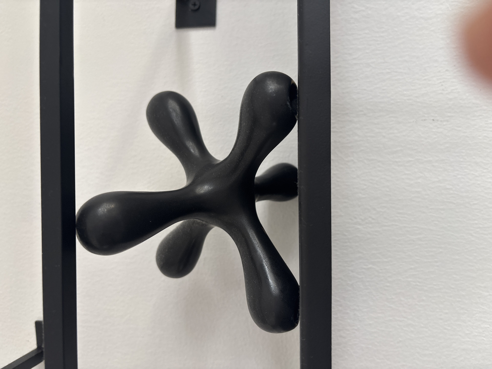 An abstract black sculpture, resembling a jack or a starburst with rounded appendages, is centrally placed on a black shelf. The shelf itself is mounted on a white, textured wall. Above this shelf, another black shelf is visible. The lighting suggests an indoor environment. There is no indication of the city of Dubai or the United Arab Emirates in the image.