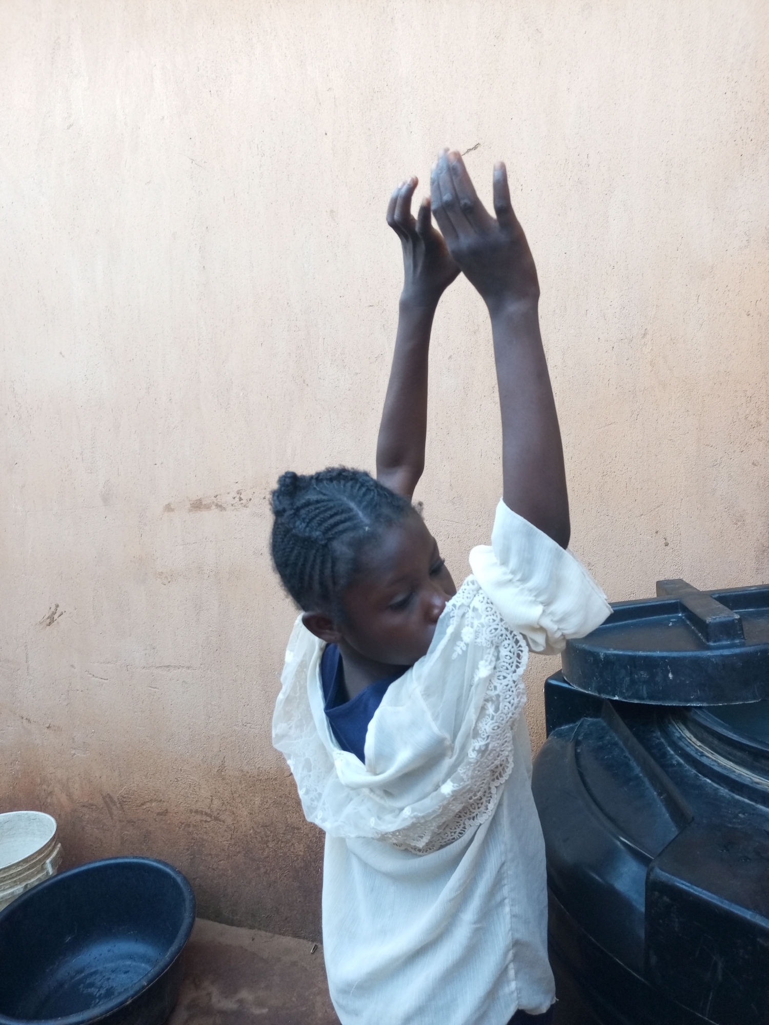 A young girl with braided hair stands in front of a textured, light-colored wall in what appears to be an outdoor area in Tempure, Nigeria. She is wearing a white dress with lace detailing on the sleeves. Her arms are raised, and her hands are held in front of her face, palms open and facing forward, as if she is clapping or preparing to catch something. To her right, a large black water tank is partially visible. In the foreground, to the left, are two open containers: a black oval basin and a white bucket. The ground appears to be dirt or concrete.