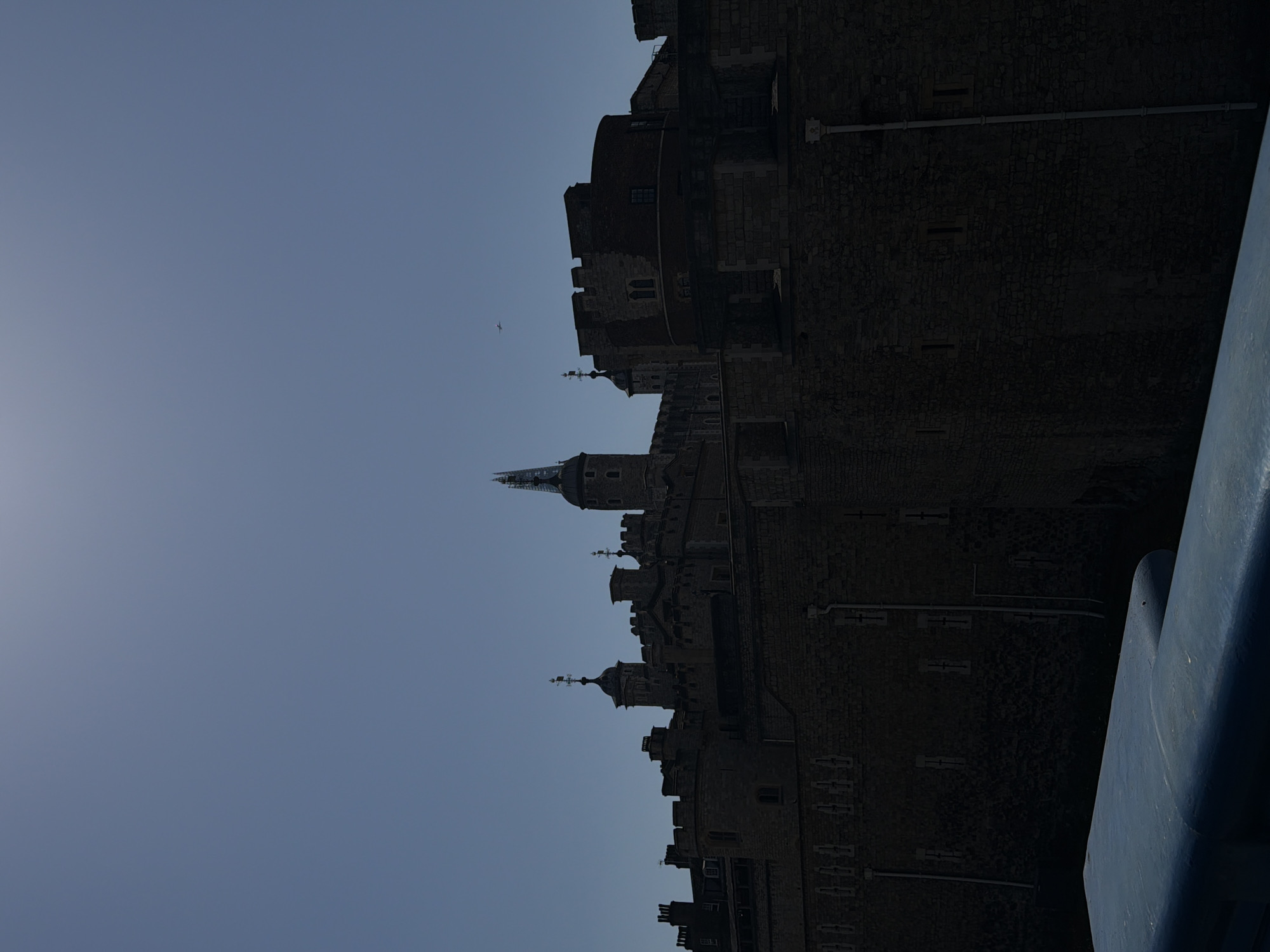 The image depicts the Tower of London in London, United Kingdom. The structure is silhouetted against a light blue sky. Architectural details include a pointed spire, crenellations, and various towers. A small bird is visible in the sky. The lower right corner of the image includes a blue structure.