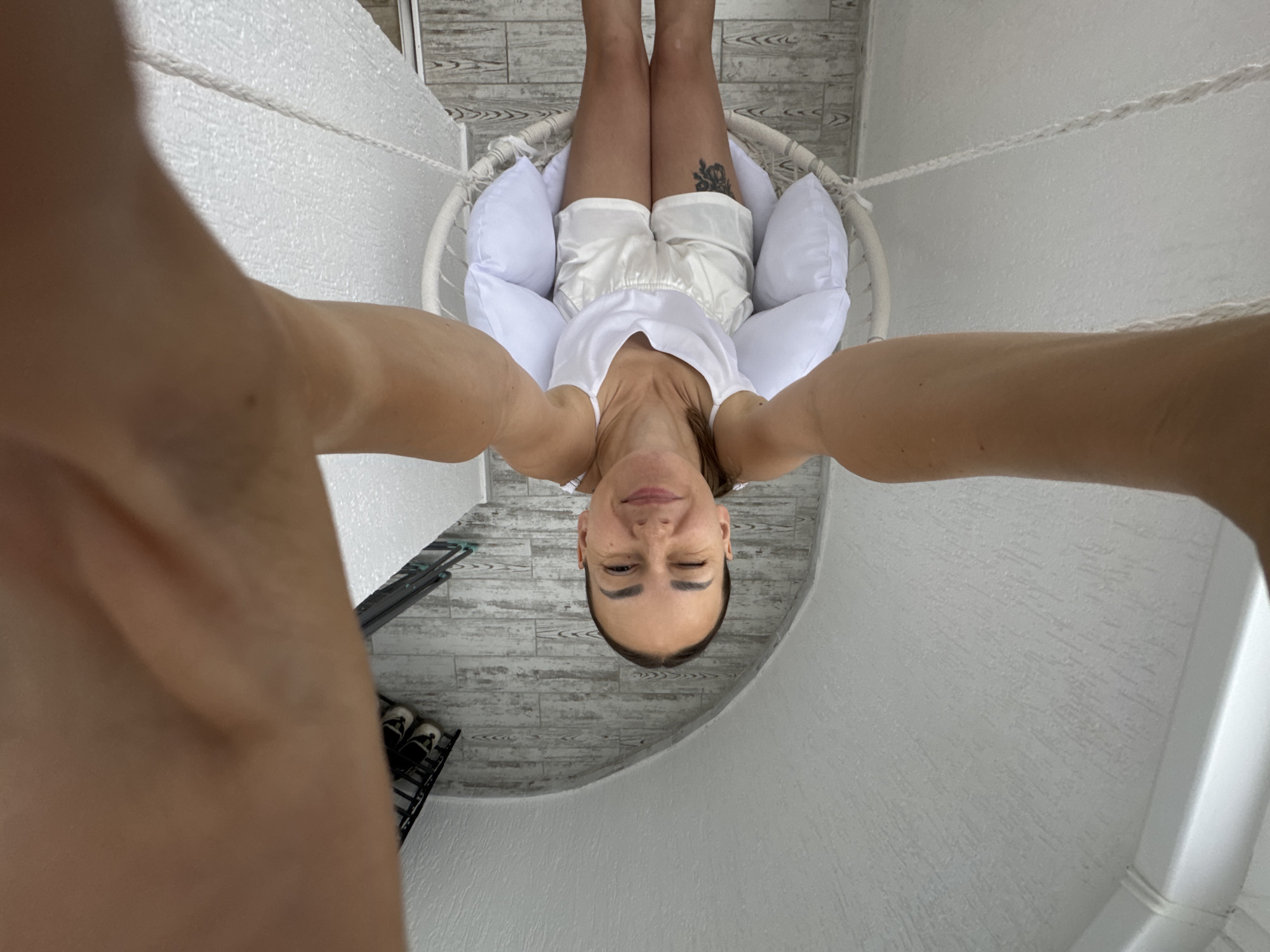 A woman is sitting in a hanging chair outdoors, possibly on a balcony or patio. She is wearing a white tank top and white shorts. She has a tattoo on her left thigh. The chair is white and suspended by ropes. There are white pillows in the chair. The flooring appears to be wood planks. In the background, there is a shoe rack with shoes on it. The scene is set in Sochi, Russia. The woman is smiling and looking at the camera. The photo is taken from a high angle.