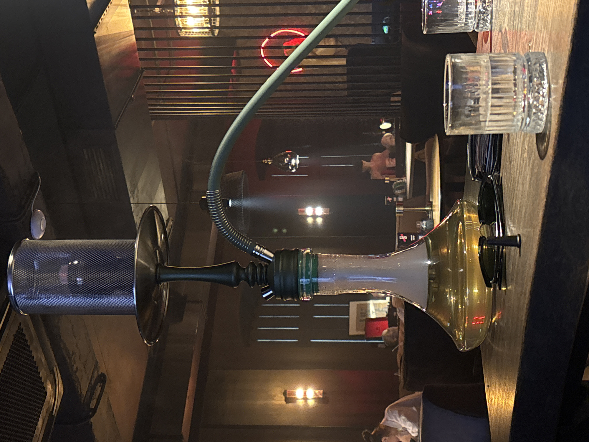 The image depicts a close-up view of a hookah setup in what appears to be a dimly lit lounge or bar. The hookah is the central focus, with its metallic top and glass base filled with a yellow liquid. A light blue hose extends from the hookah, curving out of frame. In the foreground, a wooden table holds two glasses of clear liquid, sunglasses, and a wallet or stack of cards. In the background, the environment suggests a public space with patrons and decorative lighting. The overall ambiance is one of relaxation and socializing. The location is specified as a shisha bar in the city of Kazan, Russia.
