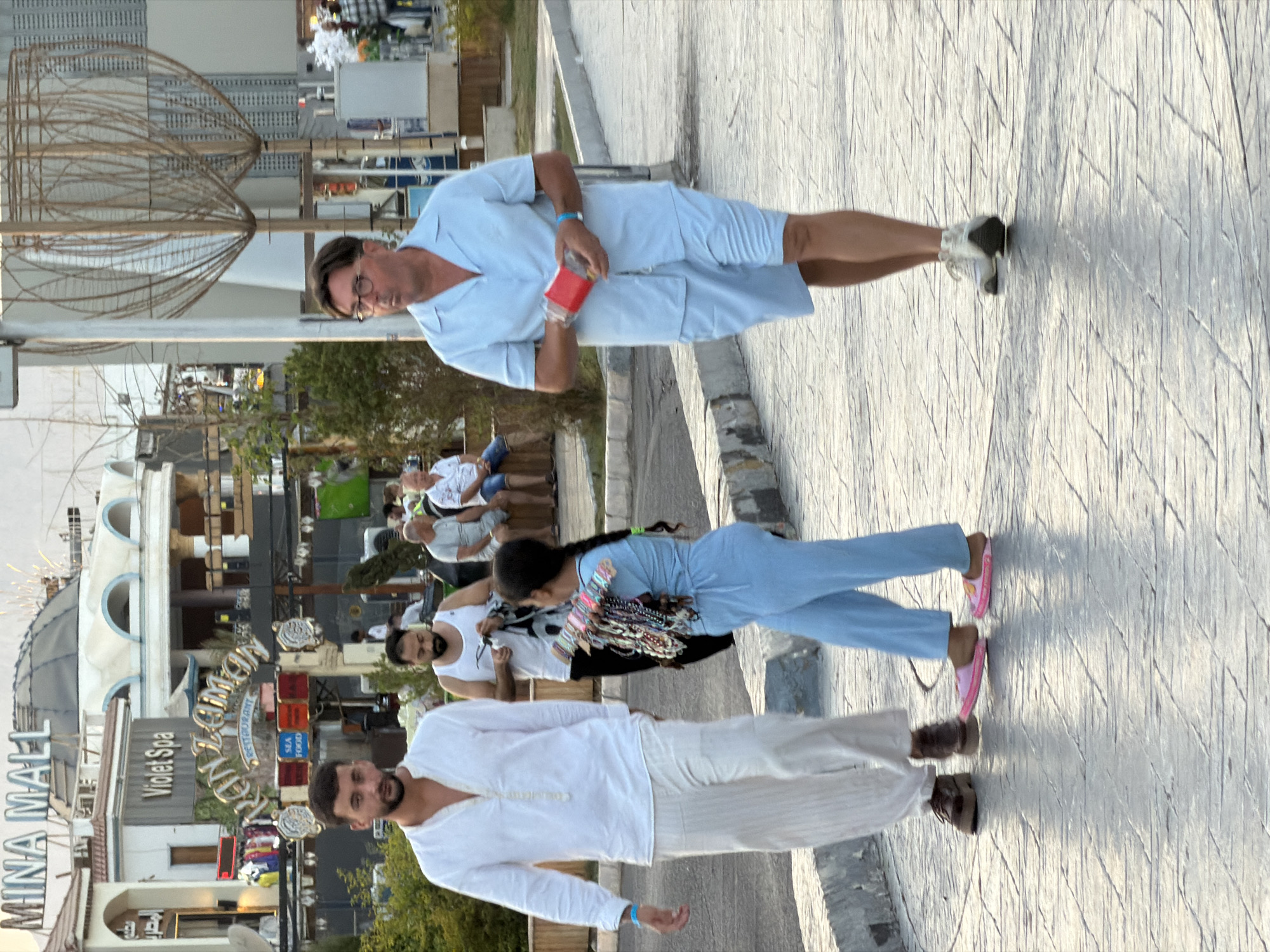 The image depicts a street scene in Sharm El Sheikh, Egypt, likely in a commercial or tourist area, possibly near "Mina Mall" as indicated by signage. In the foreground, three individuals are walking on a paved walkway.
On the left, a man in a white, loose-fitting, long-sleeved shirt with a mandarin collar and loose, off-white pants, walks towards the viewer's left. He has dark hair and a beard, and is wearing dark shoes.
To his right and slightly behind him, a young girl is walking towards the right. She has dark hair braided down her back and is wearing a light blue, long-sleeved top and matching pants. She is carrying a bundle of colorful necklaces and bracelets on her right arm, presumably for sale.
On the right side of the frame, a man wearing a light blue short-sleeved shirt with a camp collar and matching shorts walks towards the viewer's right. He has short brown hair and is wearing glasses. In his hands, he is holding a red rectangular object. He is wearing white sneakers.
In the background, there are several buildings with architectural details common in resort towns, including arched windows and domes. Signs for "Violet Spa" and "Kan Zaman Restaurant" are visible, along with a "SEA FOOD" sign. Other people are visible in the middle ground, some sitting on a bench. The environment appears to be an outdoor pedestrian area with some greenery and decorative metal structures. The lighting suggests it is daytime, likely late afternoon due to the long shadows.