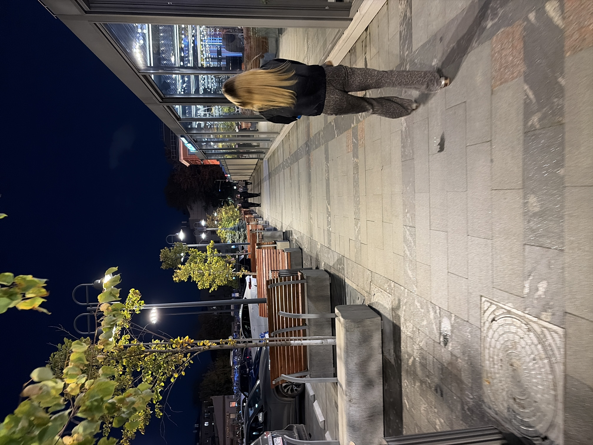 The image captures a nighttime scene on a city sidewalk, likely in Moscow, Russia, given the Cyrillic text visible on signage and car license plates. A woman with long blonde hair, wearing a dark jacket and patterned pants, walks away from the camera down a paved sidewalk lined with modern streetlights and trees. To her right, a long glass-fronted building, possibly a restaurant or shop, is illuminated from within, reflecting the interior lights and the night sky. To her left, a row of wooden benches with concrete bases sits adjacent to a parking area where cars are visible. The sidewalk is paved with large, light-colored stone tiles, and there are some wet patches, indicating recent rain. The overall atmosphere is one of urban activity continuing into the evening.