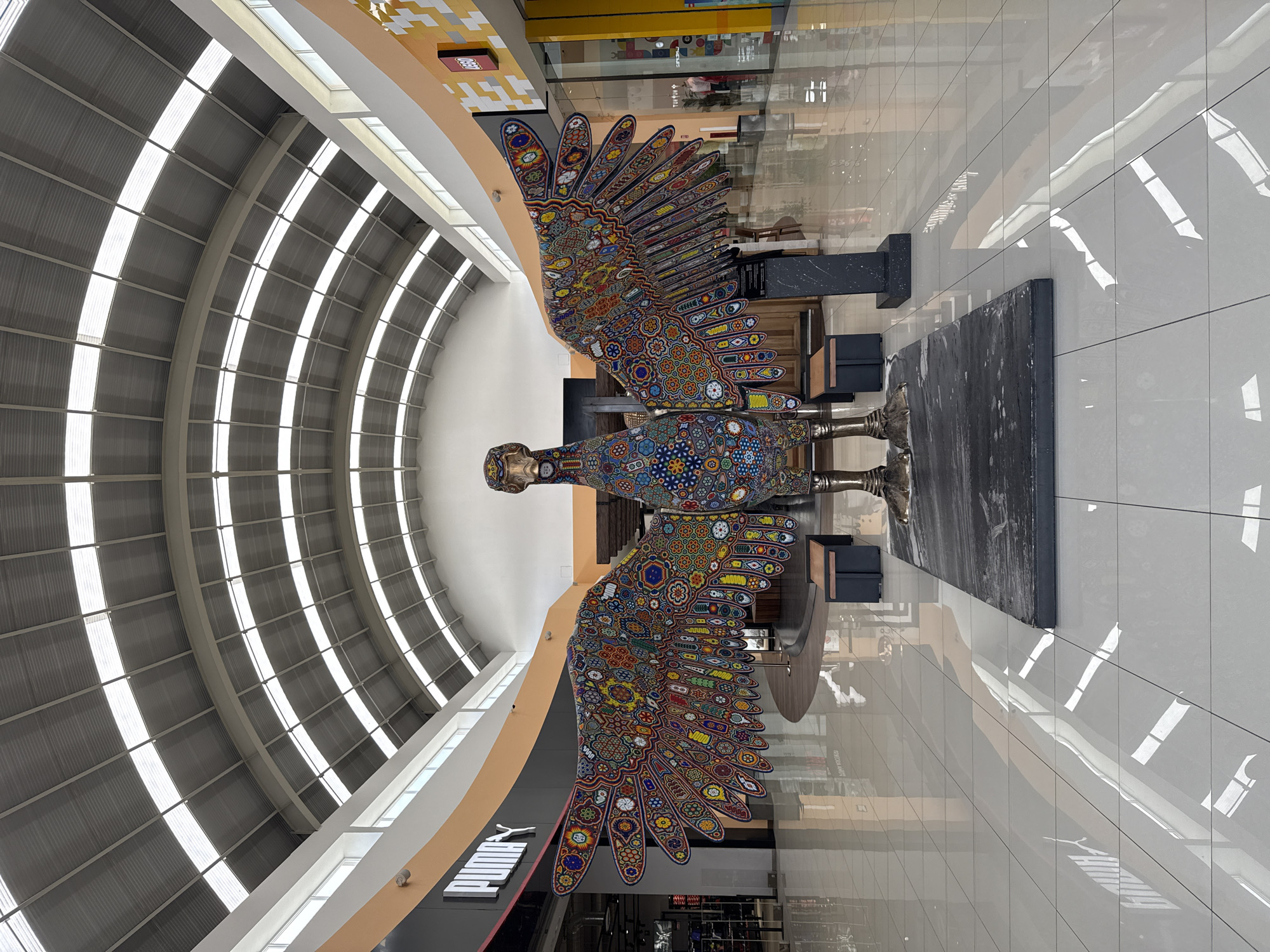 The image captures an indoor scene, likely a shopping mall or airport terminal in Ocotlán, Mexico. The focal point is a large, ornate sculpture of a winged figure, perhaps an eagle or mythical creature, adorned with colorful, intricate patterns suggestive of indigenous Mexican art styles. The sculpture stands on a raised platform in the center of a tiled walkway. The environment includes storefronts with brand names like Puma and Lego visible in the background. The architecture features a curved, skylit roof allowing natural light into the space. The reflections on the polished floor indicate a clean and well-maintained environment. No people are directly visible in the image.