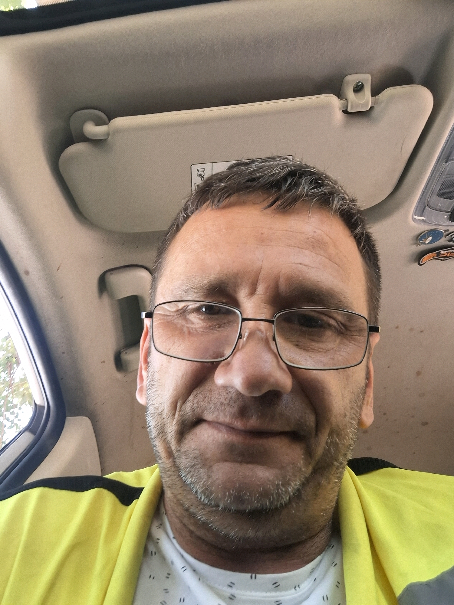 A selfie taken from inside a vehicle shows a middle-aged man with graying hair and a beard, wearing glasses and a yellow high-visibility vest over a white patterned shirt. He is looking directly at the camera. The vehicle's interior, including the sun visor and ceiling, is visible in the background. The image was captured on a sunny day, with foliage visible through the car window. The man's face is well-lit, and his expression is neutral.
