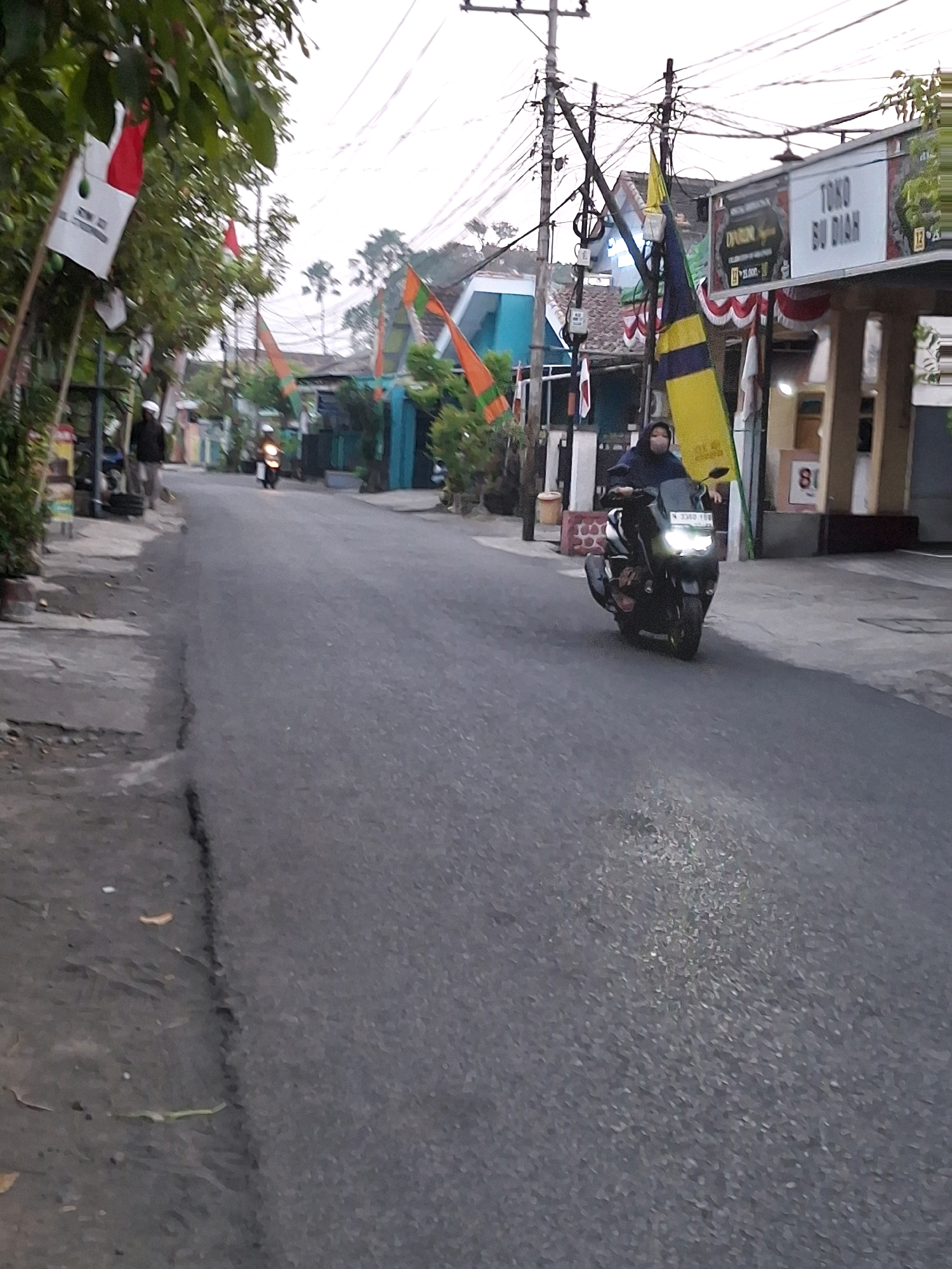 The image shows a street scene in Lumajang, Indonesia. A person is riding a scooter down a paved road. The scooter has a large yellow and blue flag attached to it. There are buildings on both sides of the road, some of which are decorated with flags. Overhead, there are power lines and poles. The sky is overcast. One building has a sign that reads "TOKO SU DIAR." A person in a dark jacket and mask is riding the scooter. Another person is standing on the sidewalk to the left of the road, wearing a white head covering and dark clothing. Trees and foliage are visible on the left side of the road, with flags hanging from some of the trees.