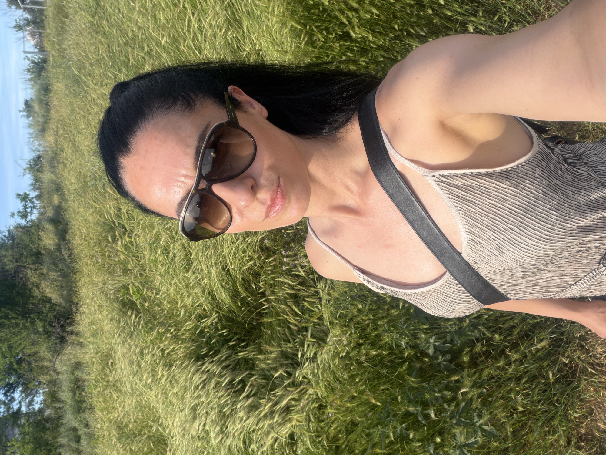 This is a selfie taken outdoors, likely in a grassy field with tall, windblown grass. The subject is a woman with long, dark hair, wearing sunglasses and a striped dress. She is lying down or leaning back in the grass. The sun is casting a warm glow, suggesting it is daytime, possibly late afternoon or early evening due to the angle of the light. The sky is visible in the background, appearing clear and blue. The overall mood is relaxed and natural, with the subject appearing comfortable in the outdoor setting. There is no visible text in the image.