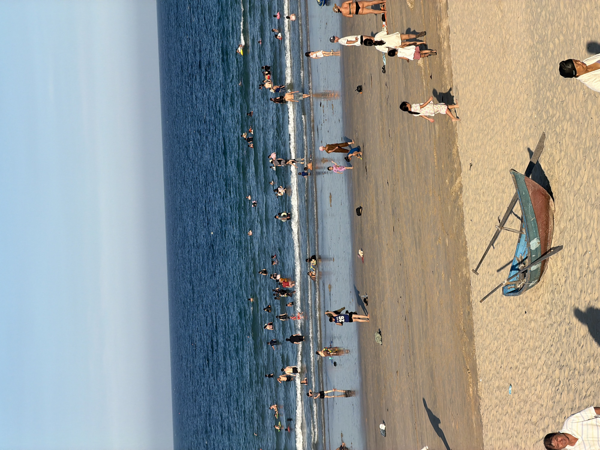 The image depicts a crowded beach scene in Cam Lam, Vietnam, likely during daylight hours given the bright sunlight and clear sky. The ocean is a deep blue, with moderate waves lapping at the shore. Numerous people are in the water, some swimming, others standing or wading. On the sandy beach, a variety of activities are taking place. Several individuals are walking or standing, while children are seen playing. A few boats, including a partially submerged blue and red one with oars, are present on the sand. Clothing, bags, and a plastic bottle are scattered across the beach. The overall atmosphere suggests a popular recreational area.
