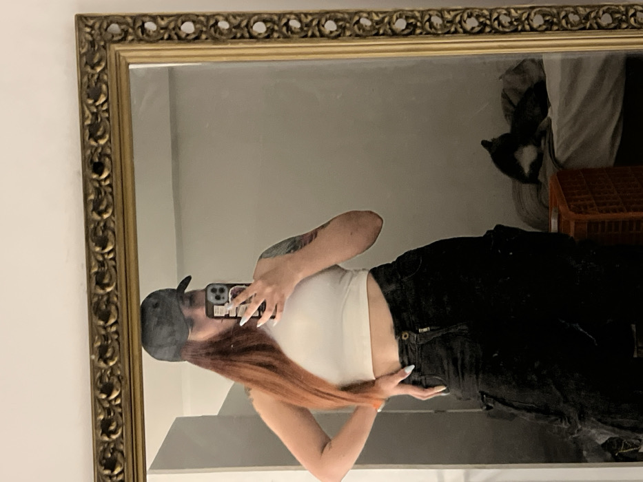 A person takes a mirror selfie. They wear a gray baseball cap, a white crop top, and black pants. Red hair is visible. The phone case is dark and has stickers on it, and the nails are long and white. The ornate gold mirror frame is clearly visible. A glimpse of a cat and an orange crate are visible in the background, along with folded white cloth. The overall image quality is slightly blurry.
