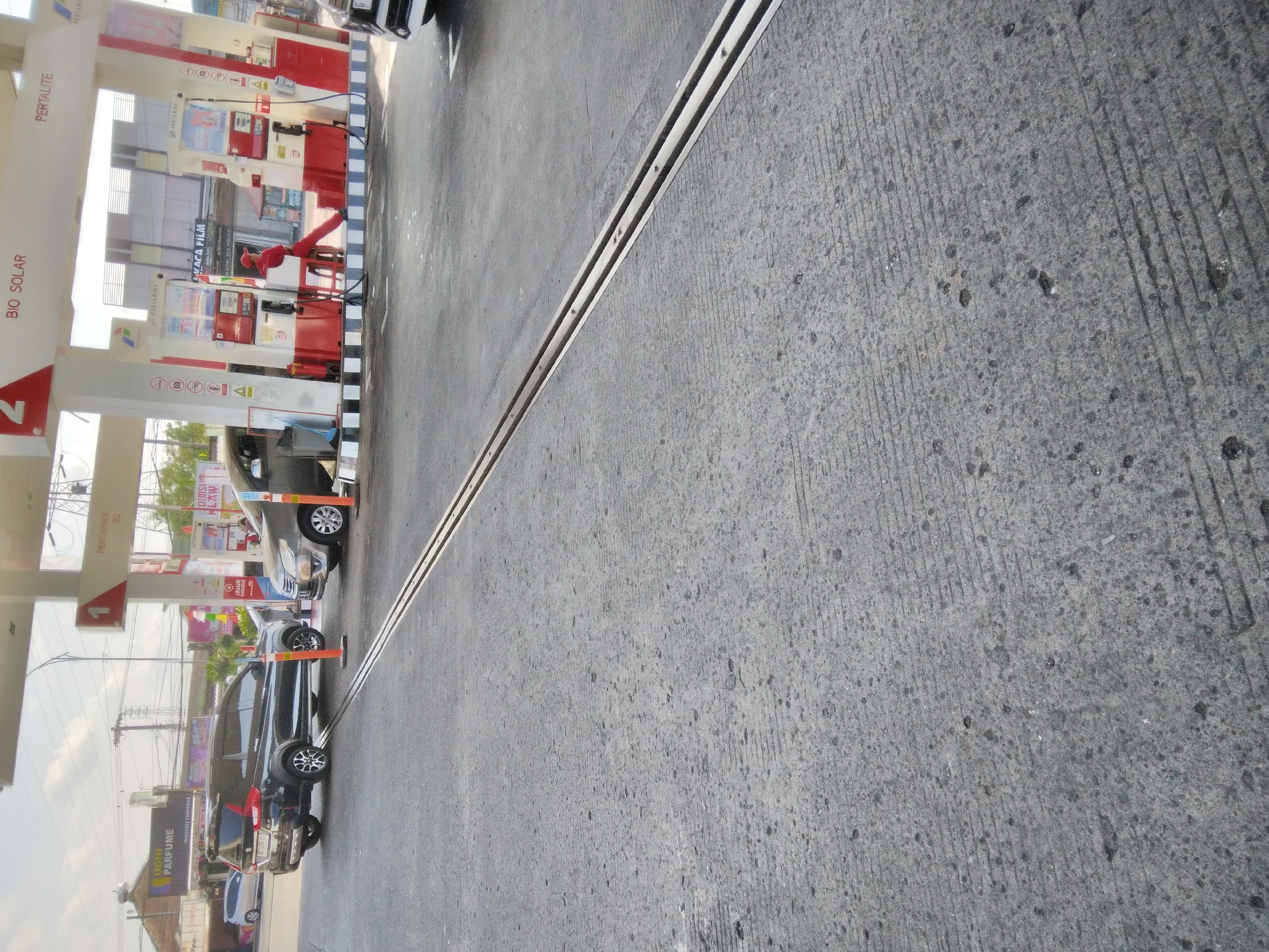 A street-level shot shows a Pertamina gas station in Purwokerto Kidul, Indonesia. The foreground is dominated by a gray asphalt road with distinct parallel lines carved into it and a metal expansion joint running diagonally across the image. In the middle ground, two vehicles are parked at a gas pump. A dark-colored SUV with a rear spoiler is at the left pump, and a silver minivan is at the right pump. An attendant wearing a red uniform and cap sits on a stool near the pumps, facing away from the camera. The gas station canopy is visible above, with Pertamina branding and fuel type labels such as "BIO SOLAR" and "PERTALITE." Signs indicating "PERTAMINA" are mounted on the white support pillars of the canopy. In the background, buildings and more signage are visible. The sky is overcast.