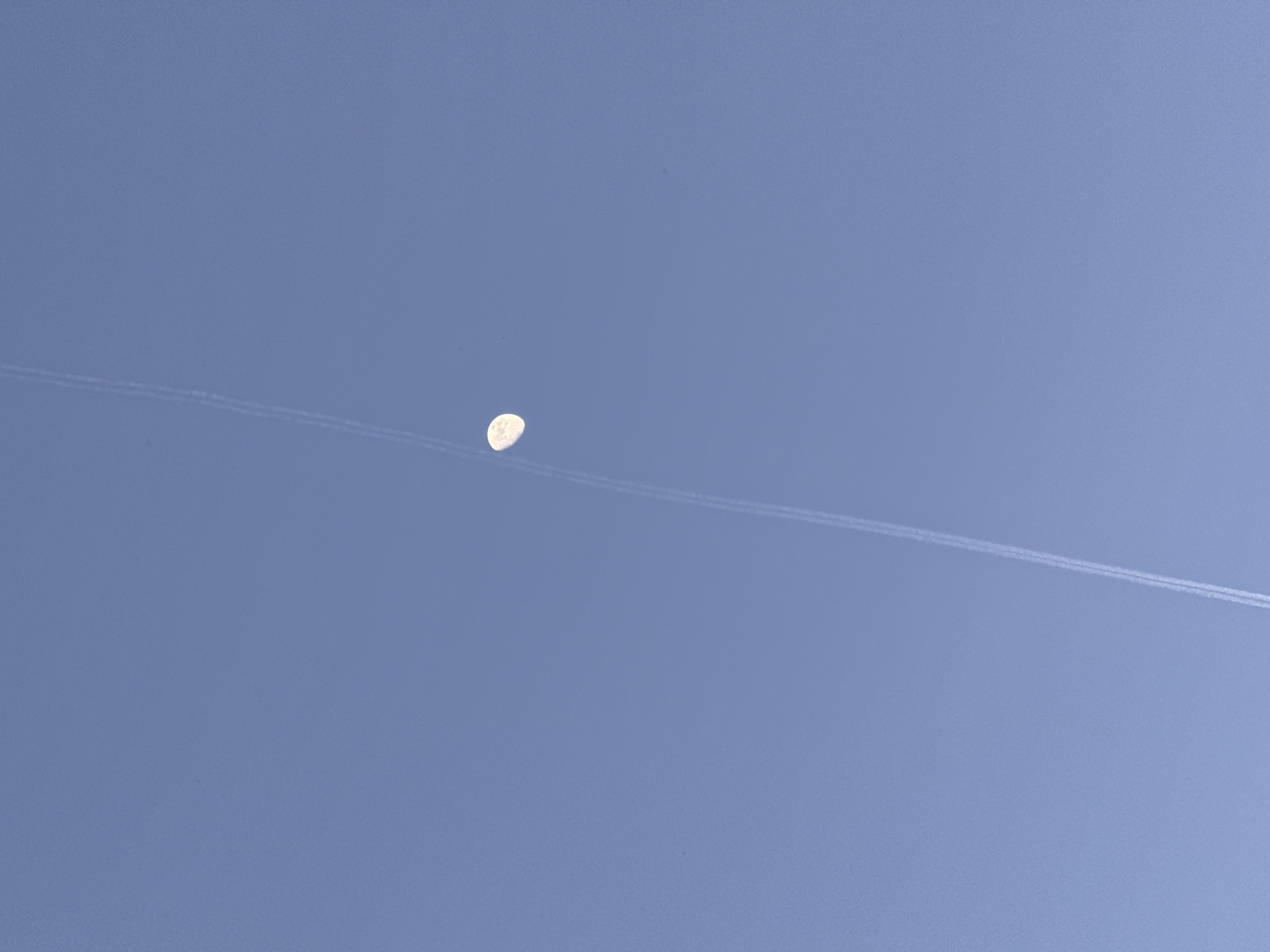 The image shows a clear blue sky with a contrail running horizontally across the frame. In the center, there is a waning gibbous moon. The contrail appears to pass behind the moon. There are no people visible, and the location is not discernable as Belgrade, Serbia.