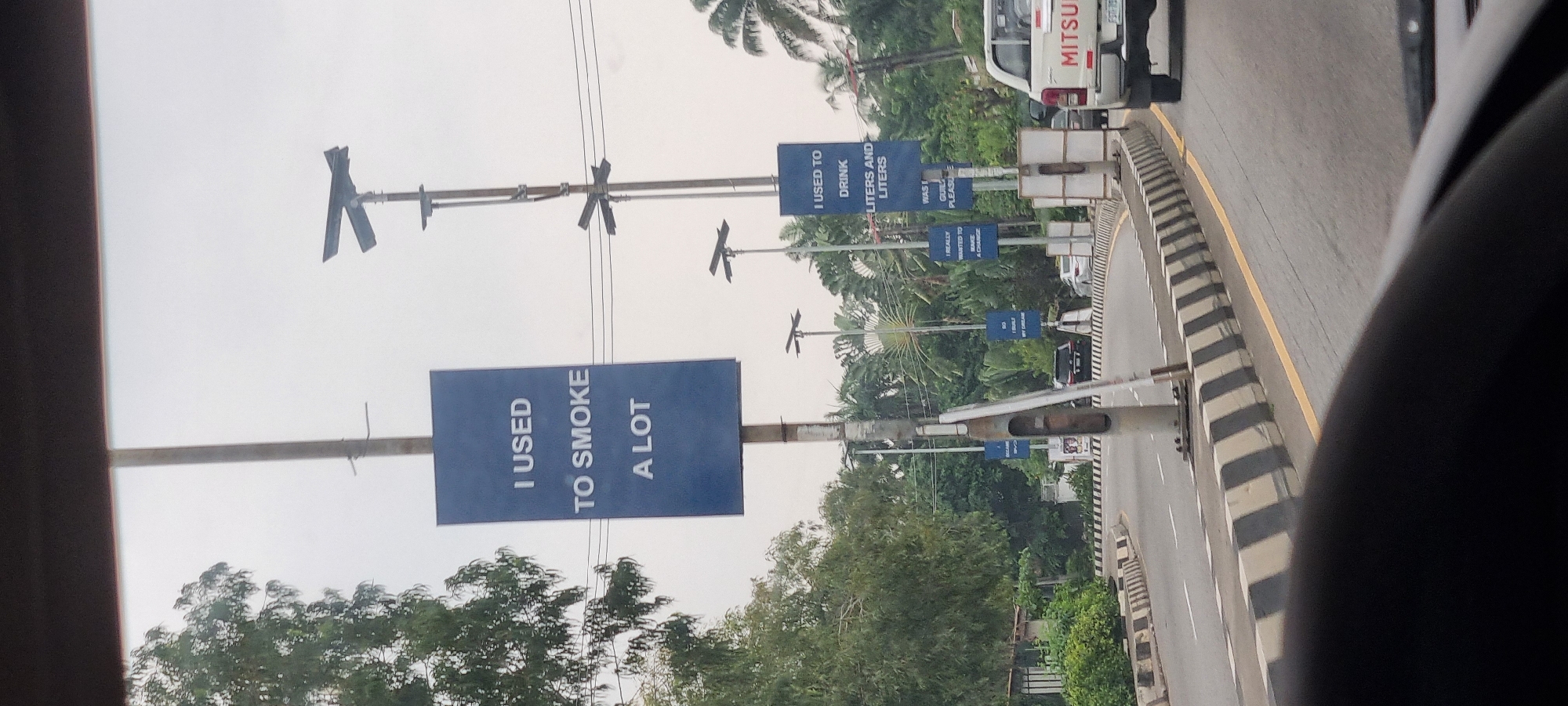 This is a street-level, eye-level, outdoor, daytime, medium shot taken from a vehicle, looking forward. The image captures a road in Ikoyi, Nigeria, with a prominent set of blue billboards on light poles. The billboards convey personal affirmations and past habits. The first billboard on the left reads "I USED TO SMOKE A LOT." Further down the road, another billboard states, "I USED TO DRINK LITERS AND LITERS." Additional, partially visible billboards suggest themes of change and aspiration, with phrases like "I REALLY WANTED TO MAKE A CHANGE" and "SO I BUILT MY DREAM." The road itself is paved and curves to the right, with clear lane markings and black-and-white striped traffic barriers. Lush green trees and palm trees line the sides of the road, indicating a tropical environment. A white Mitsubishi pickup truck is visible in the right lane, as are other vehicles in the background. The sky is overcast, suggesting a cloudy or hazy day. The overall scene depicts a public space in a Nigerian city adorned with motivational messaging.