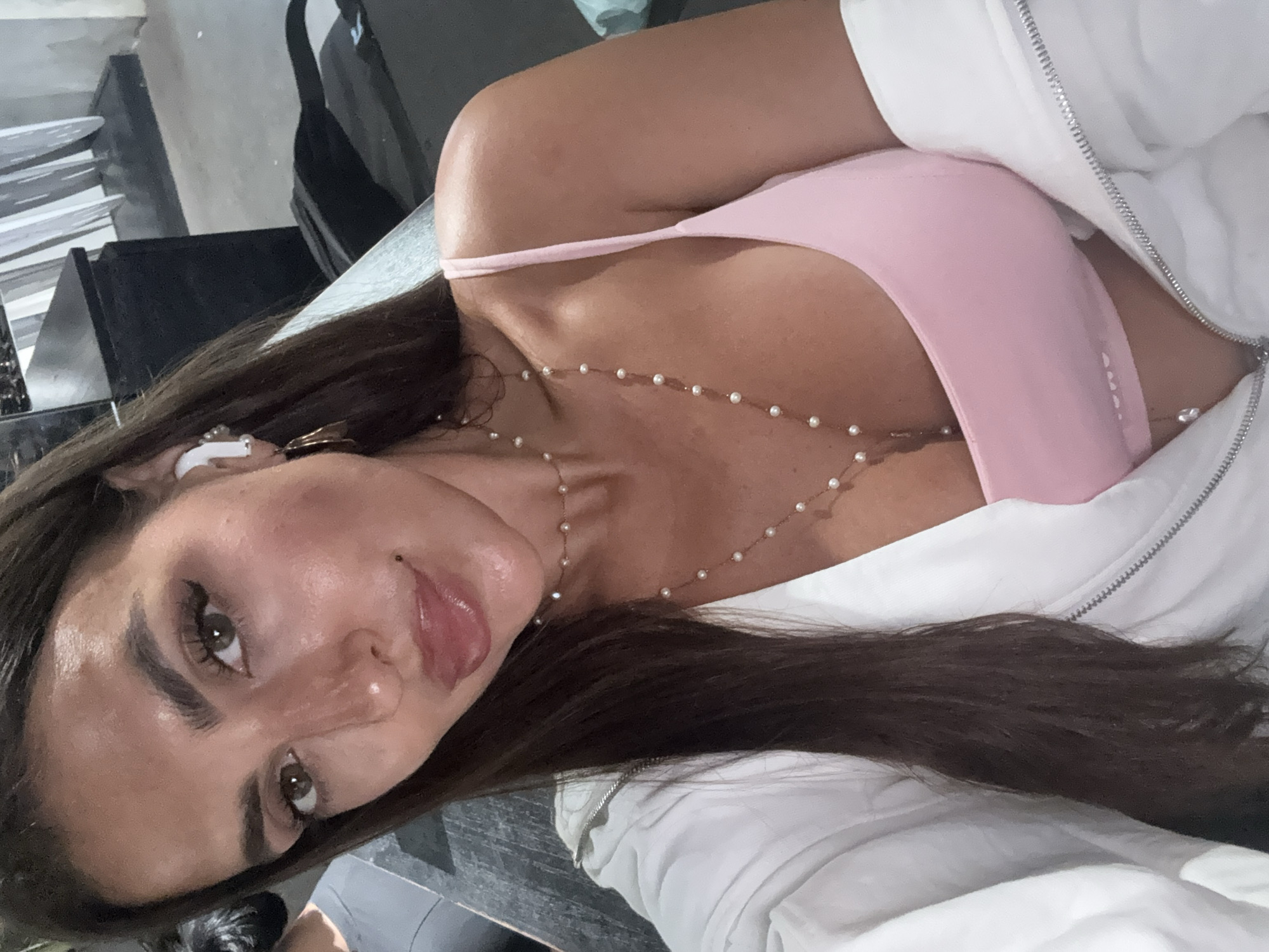 A medium close-up shot presents a woman with dark hair and light skin, positioned at an oblique angle. She is wearing a light pink top and a white zip-up jacket. A delicate pearl necklace adorns her neck. An earbud is visible in her ear. The background suggests an indoor setting. The image's origin is potentially North Kuta, Indonesia.