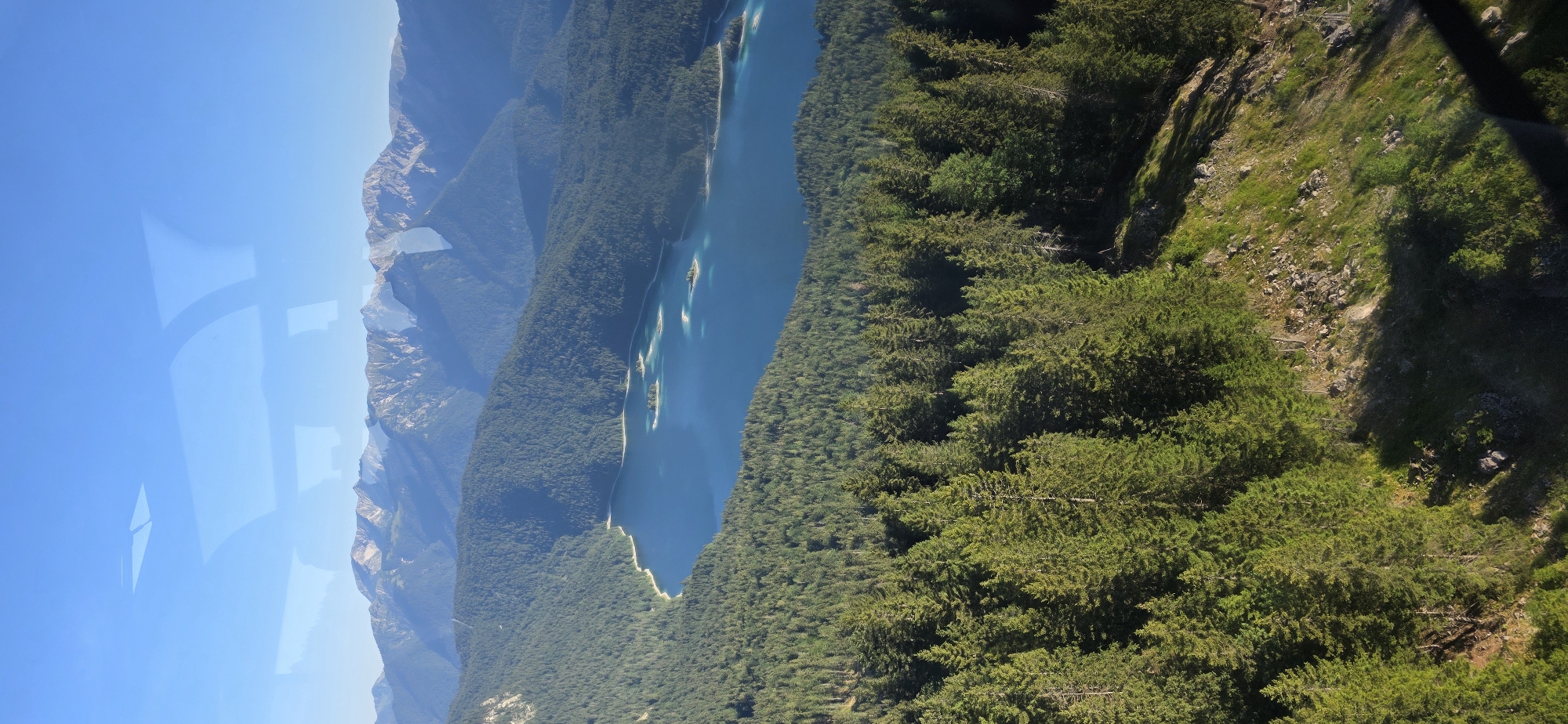 This image captures a panoramic view of the Eibsee lake in Grainau, Germany, with the Wetterstein mountain range in the background. The scene is bathed in clear, bright sunlight, casting distinct shadows across the landscape.
In the foreground, a dense forest of evergreen trees, primarily firs, blankets a steep, rocky slope. The trees are vibrant green, with visible shadows indicating the direction of the sun. The terrain is uneven, with scattered rocks and patches of grass visible between the trees.
Beyond the forest, the Eibsee lake stretches out, its deep blue water dotted with several small islands. The water's surface is calm, with subtle variations in color suggesting varying depths and underwater topography. The islands are covered in lush green vegetation.
The middle ground features rolling, forested hills that lead up to the majestic Wetterstein mountains. These mountains, with their rugged peaks and rocky faces, form a dramatic backdrop. The mountain slopes are a mix of dark green forests and lighter, rocky outcrops. The distant peaks are softened by atmospheric haze, giving them a bluish tint.
The sky above is a clear, vibrant blue, with no clouds visible. There are reflections on what appears to be a window or glass surface, distorting parts of the sky and mountain range. These reflections show geometric shapes and curved lines, possibly from the structure of the viewing point.
There are no people or animals visible in the scene. The overall impression is one of natural beauty and tranquility, showcasing a pristine mountain lake and its surrounding wilderness.